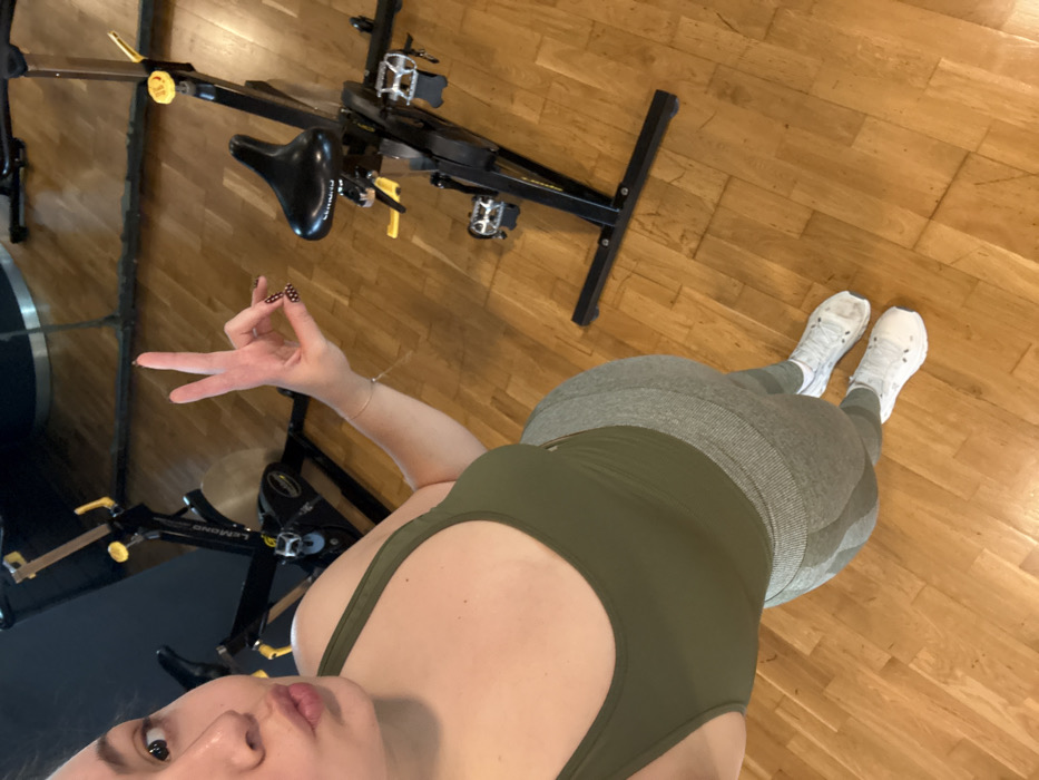 A female individual is visible in what appears to be an indoor fitness facility, specifically a spin studio. The specific location within Mexico City, Mexico, is not identifiable from the visual information provided.
The individual, seen from a high-angle selfie perspective, occupies the left and central portions of the frame. Her head is tilted, and her lips are puckered. Her right hand is raised, displaying a gesture where the thumb and index finger form a closed circle, with the remaining three fingers extended upwards. Her fingernails are painted dark with white polka dots. She is wearing a solid olive green sports top and light olive green, textured athletic leggings. White athletic socks and white athletic shoes complete her attire. A thin, light-colored bracelet is visible on her right wrist.
In the background, at least two black stationary bicycles (spin bikes) are present on a polished wooden floor. One bike in the mid-ground, labeled "LEMOND," features yellow adjustment knobs and a black seat. A dark wall with a reflective surface, likely a mirror, is visible behind the bikes. The environment suggests an indoor, maintained space.