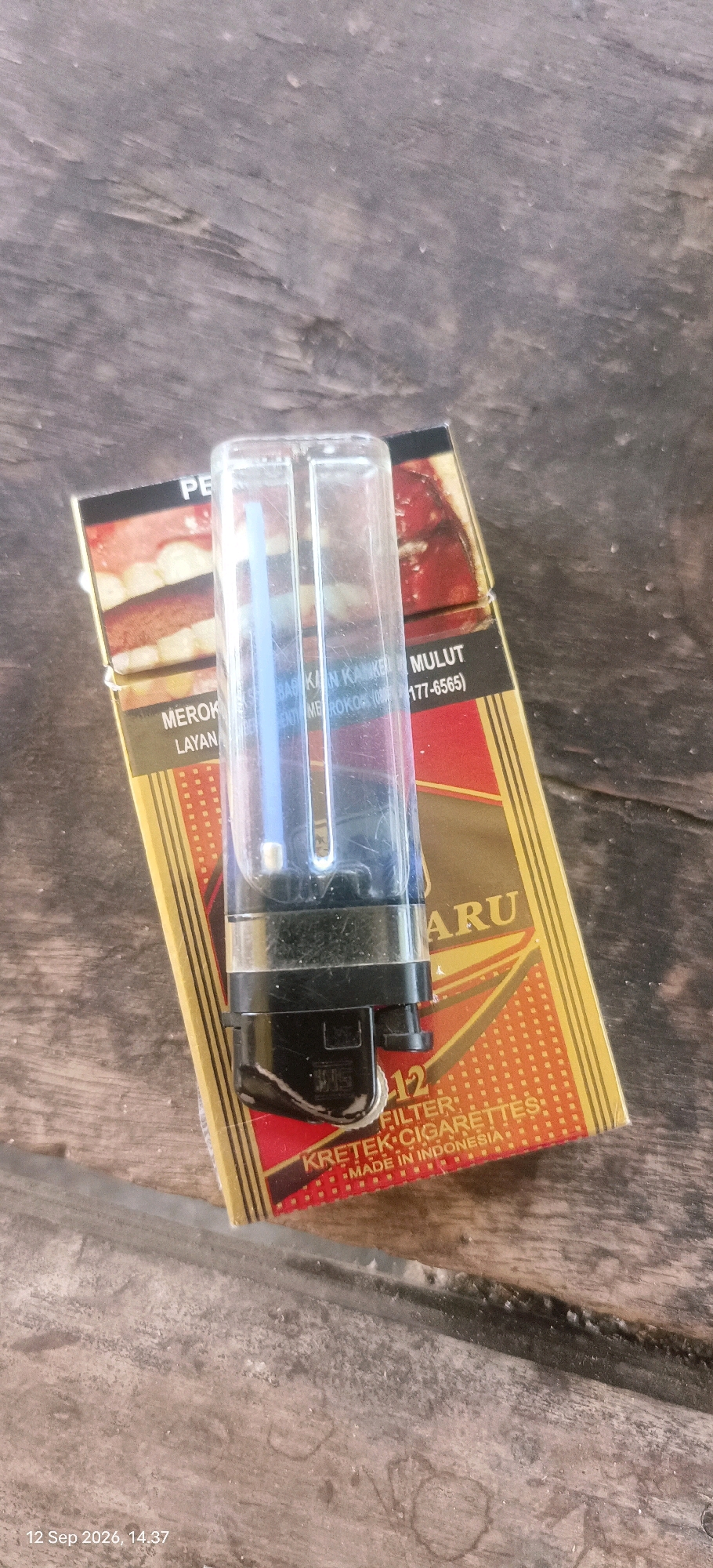 A clear plastic lighter with a blue flame mechanism sits atop a pack of cigarettes. The cigarette pack is yellow with red and gold accents and features text indicating it is a "FILTER KRETEK CIGARETTES MADE IN INDONESIA." The background is a weathered wooden surface. The timestamp "12 Sep 2026, 14:37" is visible in the lower left corner, suggesting the image was taken on that date and time. The scene is likely an outdoor setting, given the lighting and texture of the wooden surface, possibly a table or bench in Mojopetung, Indonesia. There are no people or discernible actions occurring in the image.