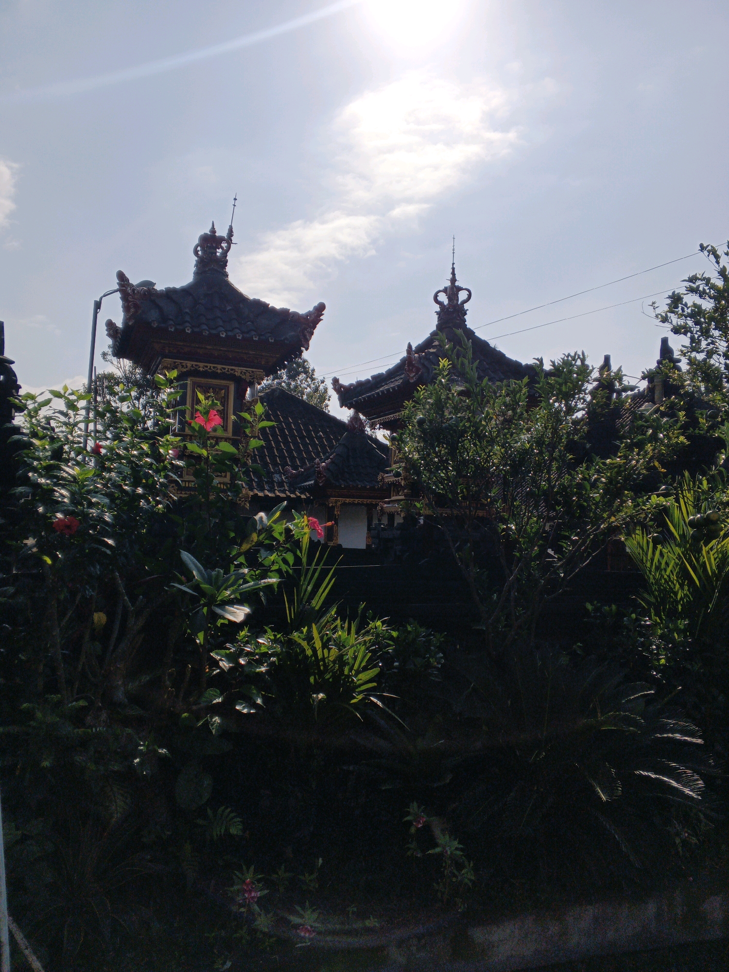 The photograph shows the exterior of a building with traditional Balinese architecture, characterized by tiered, dark tiled roofs adorned with decorative elements and finials. Two distinct structures with these roofs are visible, separated by dense green foliage. The building appears to be set within a garden or natural environment, with vibrant green plants, bushes, and some flowering plants, including a bright red hibiscus, in the foreground and surrounding the structures. The sky above is a bright, hazy blue with scattered clouds, and a lens flare from the sun is prominent in the upper left quadrant, indicating a sunny day. The overall impression is of a serene, possibly religious or cultural site, viewed from a lower vantage point, partially obscured by vegetation. This location is identified as being in the city of Penebel, Indonesia.