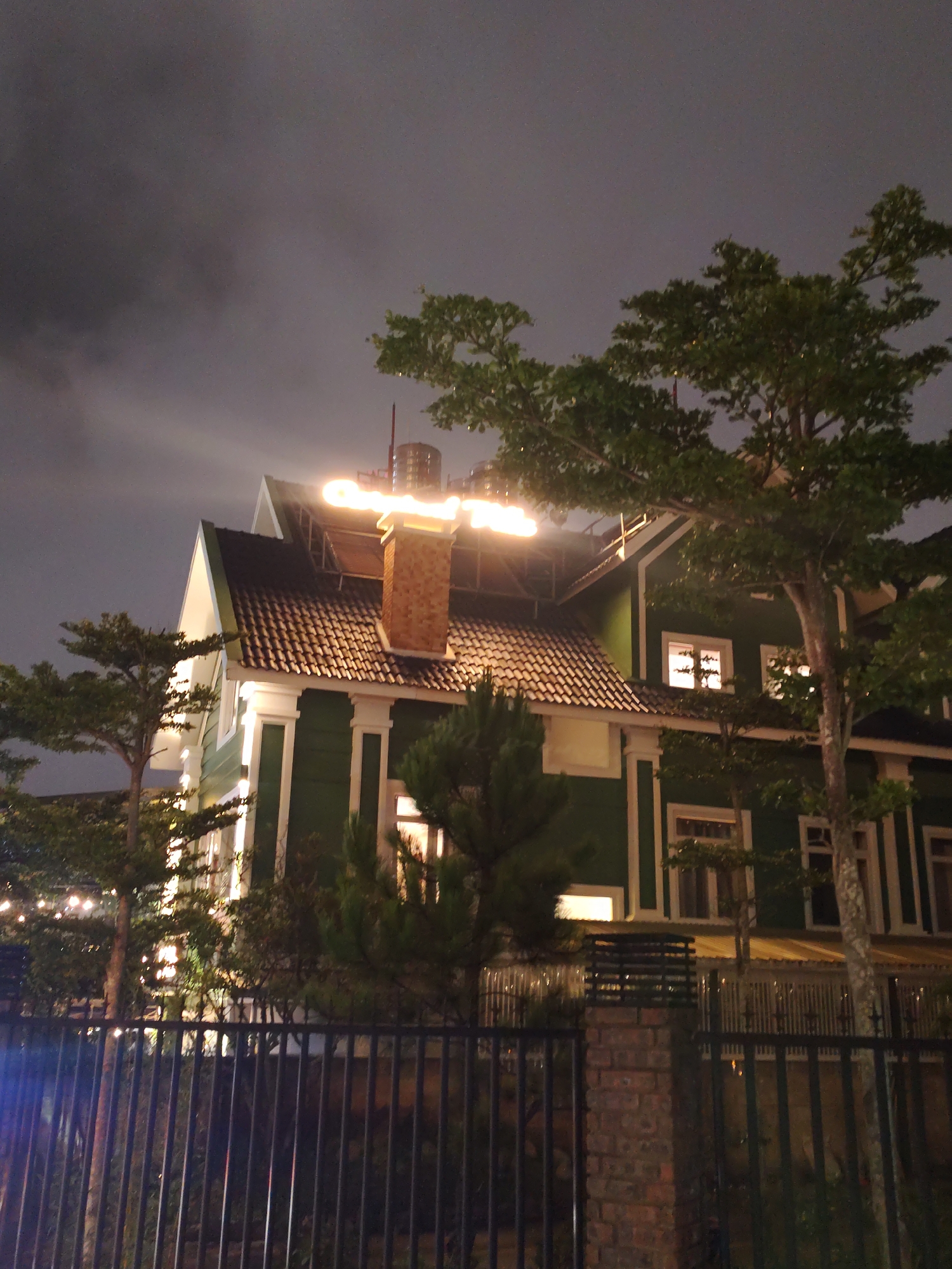 The image displays a large, two-story house at night, illuminated against a dark, overcast sky. The house is characterized by dark green siding, prominent white trim, and a steep, gabled roof covered with reddish-brown tiles. A tall, brick chimney stands centrally on the roof.
Bright, warm-toned lights are extensively used, particularly on the upper levels of the house and around the roofline, creating a striking glow against the dark environment. Above and to the right of the chimney, an illuminated sign is clearly visible, spelling out the word "GARDEN." Several windows on the house's facade also emit a warm light from within.
The property is surrounded by dense greenery, including several tall trees with dark foliage and smaller, bushy evergreen trees that partially obscure the house. In the foreground, a sturdy black metal fence runs across the bottom of the frame, accompanied by a brick pillar on the right. Behind the fence, on the left, subtle string lights are visible, contributing to the ambient nighttime illumination.
The scene depicts a static view of the building, with no people or specific activities taking place. The time of day is clearly night, evidenced by the dark, cloudy sky and the reliance on artificial lighting. The atmosphere is quiet and somewhat serene, suggesting a resort or a large, well-maintained residence. The location context indicates this is in Da Lat, Vietnam.