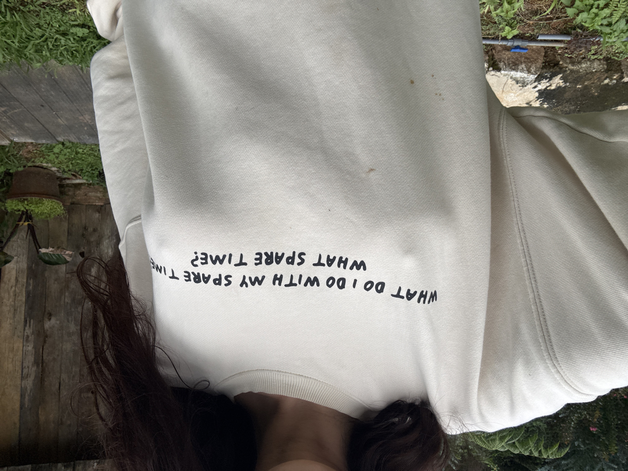 A person, only their torso visible, wears a light-colored crewneck sweatshirt with black text that reads, "WHAT DO I DO WITH MY SPARE TIME? WHAT SPARE TIME?". The person stands outdoors in what appears to be a garden setting. In the background, there's a wooden fence, green foliage, and a potted plant. The ground is covered with grass and some wooden planks. The image seems to be taken in a temperate climate. No specific landmarks of Da Lat, Vietnam, are discernible.