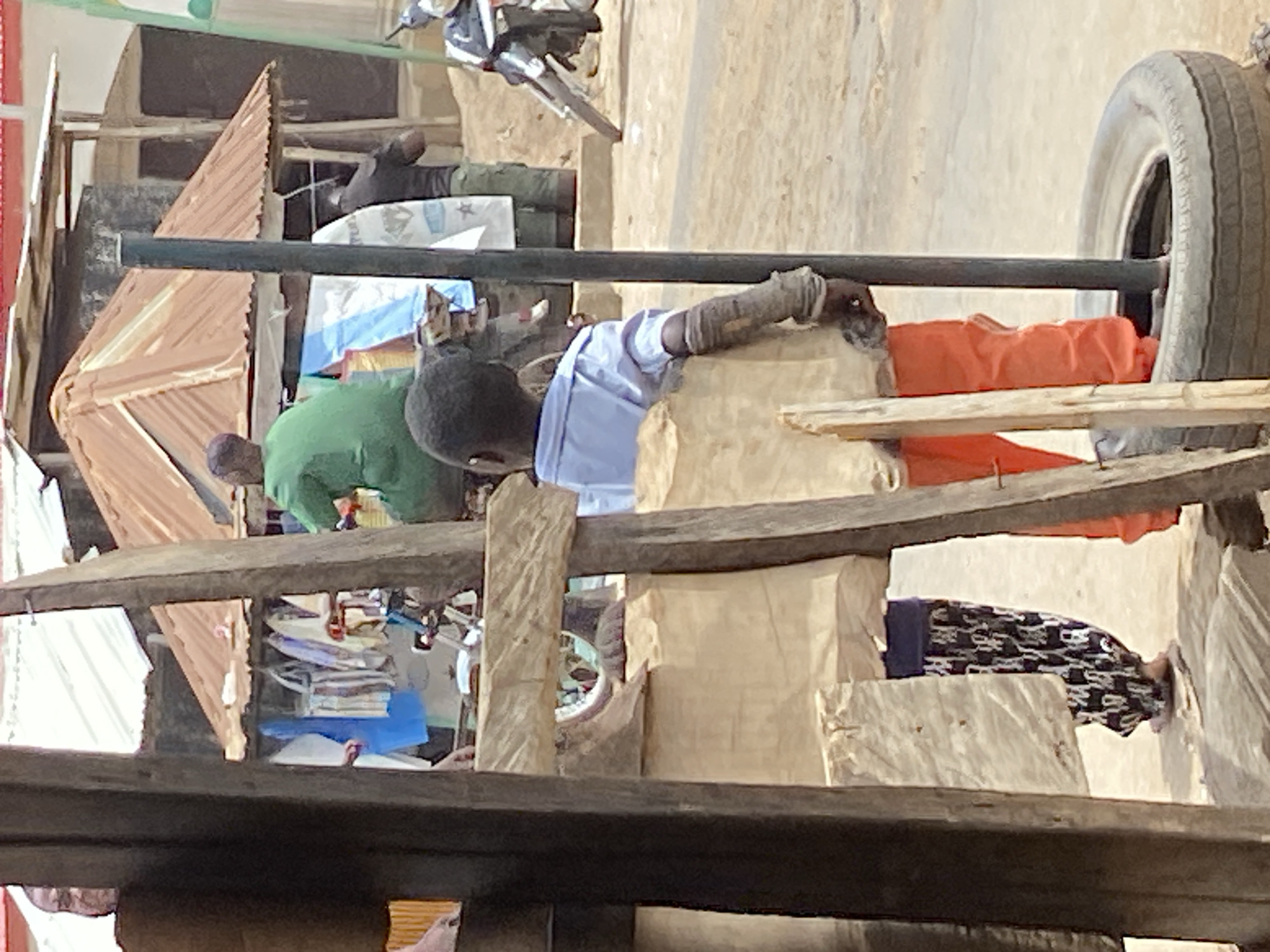 This image captures a bustling street or market scene in Mararaba, Nigeria, under bright daylight. The scene is characterized by informal structures and daily commerce.
In the foreground and midground, a young man or boy with dark skin and short hair is prominently featured, wearing a light blue short-sleeved shirt and orange pants. His left forearm is wrapped in what appears to be bandages or protective material as he leans on a dark horizontal metal pole.
To the left, slightly behind the pole, another person in a green short-sleeved shirt is seen from behind, bending over a makeshift stall or table. Further back and to the right, a person, possibly female, is partially visible, wearing a long patterned skirt (black and white/grey print).
The setting features various structures: crude wooden planks and rough-hewn timber form stalls or shelves in the foreground. In the background to the left, a stall with a reddish-brown corrugated metal roof is visible, suggesting a small shop. To the right, a light-colored wall, possibly plastered, forms the backdrop. A large vehicle tire is propped up on the far right. Parts of a motorcycle are visible in the upper background, indicating the presence of traffic. Scattered around are various goods and clutter, including stacks of items on a shelf behind the person in green and an orange piece of fabric near the tire. Several bags or sacks in the background show indistinct white patterns or writing, too blurry to discern specific words.
The activity depicted is that of daily market life, with individuals engaged in work, waiting, or general presence within a commercial area. The overall atmosphere suggests a busy, functional street environment. The light indicates it is daytime, and the weather appears clear and dry.