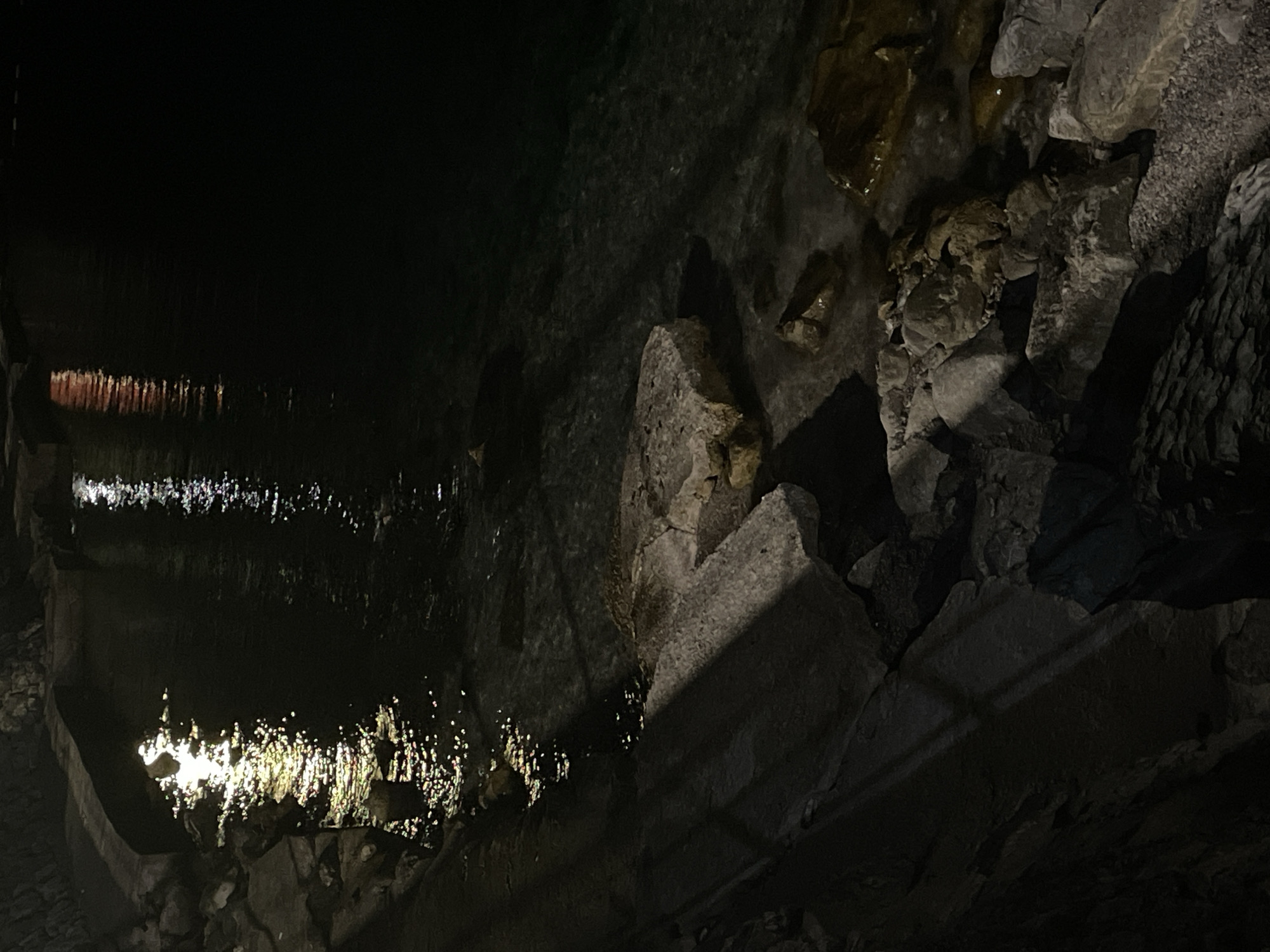 The image depicts a nighttime scene at the waterfront in Herceg Novi, Montenegro. The foreground is dominated by a rocky shoreline, with large stones and pebbles visible. The water is dark and appears relatively calm, with reflections of lights from an unseen source creating shimmering patterns on its surface. To the left, there is a curved, paved walkway or wall along the edge of the water. The lighting is dim, suggesting it is late at night or in a poorly lit area. There are no people or animals visible in the frame. The environment appears to be a coastal area within the city.