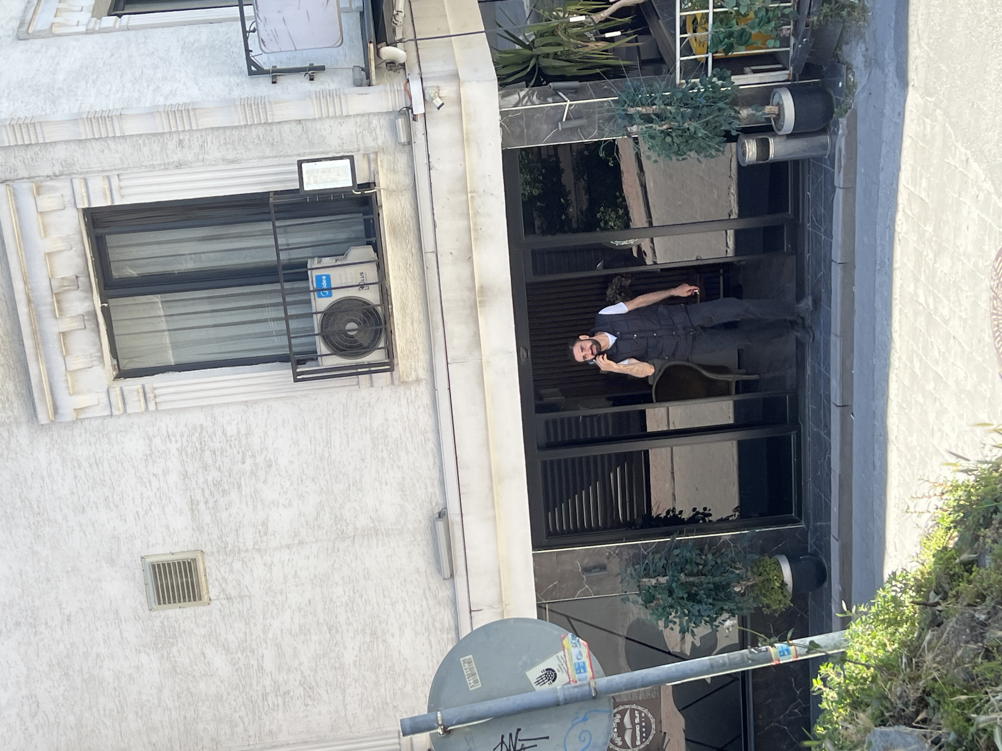 The image shows a building facade in Şişli, Turkey. The building is light-colored, with a window protected by a metal grate and an air conditioning unit. A man is standing in the building's entryway, talking on a cell phone. The entryway has a dark, reflective surface and is decorated with plants in pots. A street sign with graffiti is visible in the foreground. The street is to the right of the building, and vegetation grows along the edge of the street.