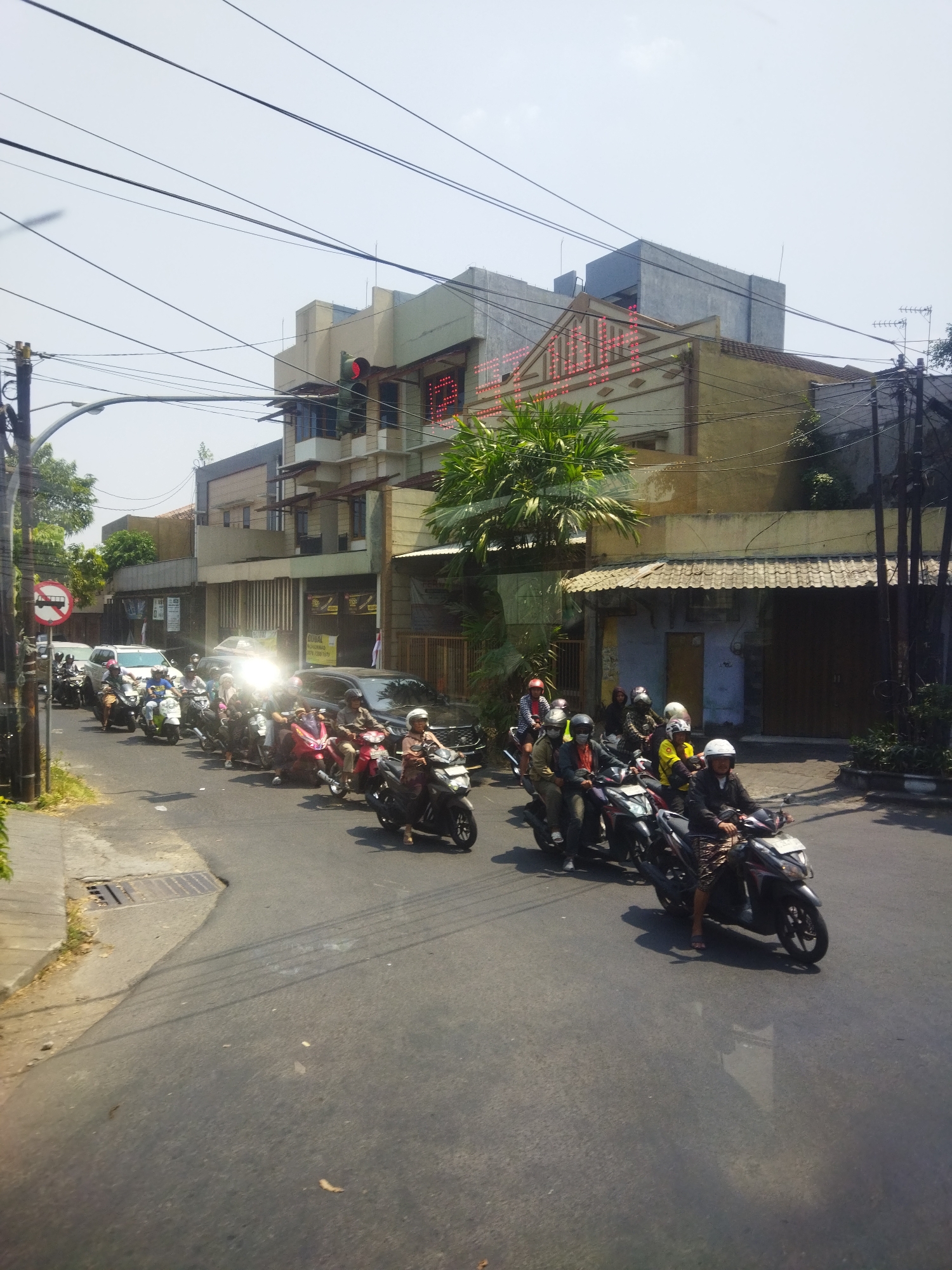 The image depicts a street scene in Surakarta, Indonesia, with a significant number of motorcycles forming a traffic jam. The foreground shows a paved road with several motorcycles, most carrying two passengers, moving slowly or stopped. One motorcycle in the immediate foreground has a rider wearing a patterned skirt and a dark jacket. Other motorcycles in view are of various colors, including red and black, with riders and passengers wearing helmets. Several cars are also present in the traffic jam, including a white car and a dark-colored car. The background features multi-story buildings, one of which has a sign with red illuminated lettering. A traffic light with a red signal is visible atop a pole. Overhead, a network of electrical wires crisscrosses the sky. The scene suggests a busy urban environment with typical traffic conditions in Indonesia.