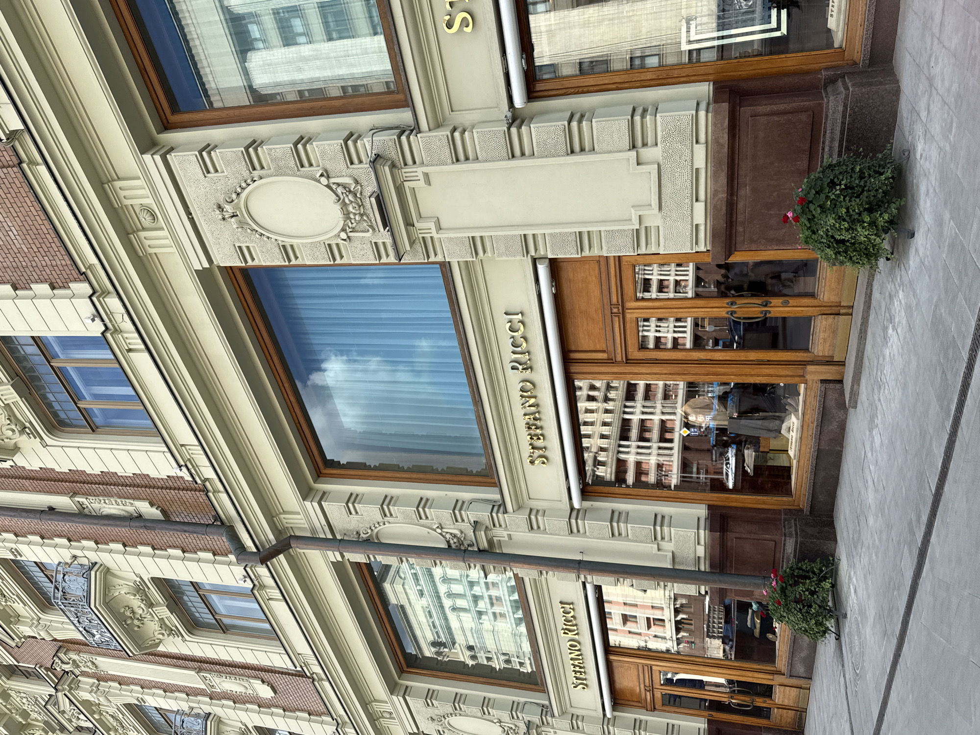 The image depicts the facade of a luxury clothing store, Stefano Ricci, located on a street in Moscow, Russia. The building features ornate architectural details with cream-colored stone accents, red brickwork, and large windows. The storefront has large wooden double doors and expansive display windows showcasing merchandise. Reflections of surrounding buildings are visible in the windows. Two decorative planters with red flowers are placed outside the entrance. The sidewalk is paved with gray stone. The overall scene is a well-maintained urban streetscape.
