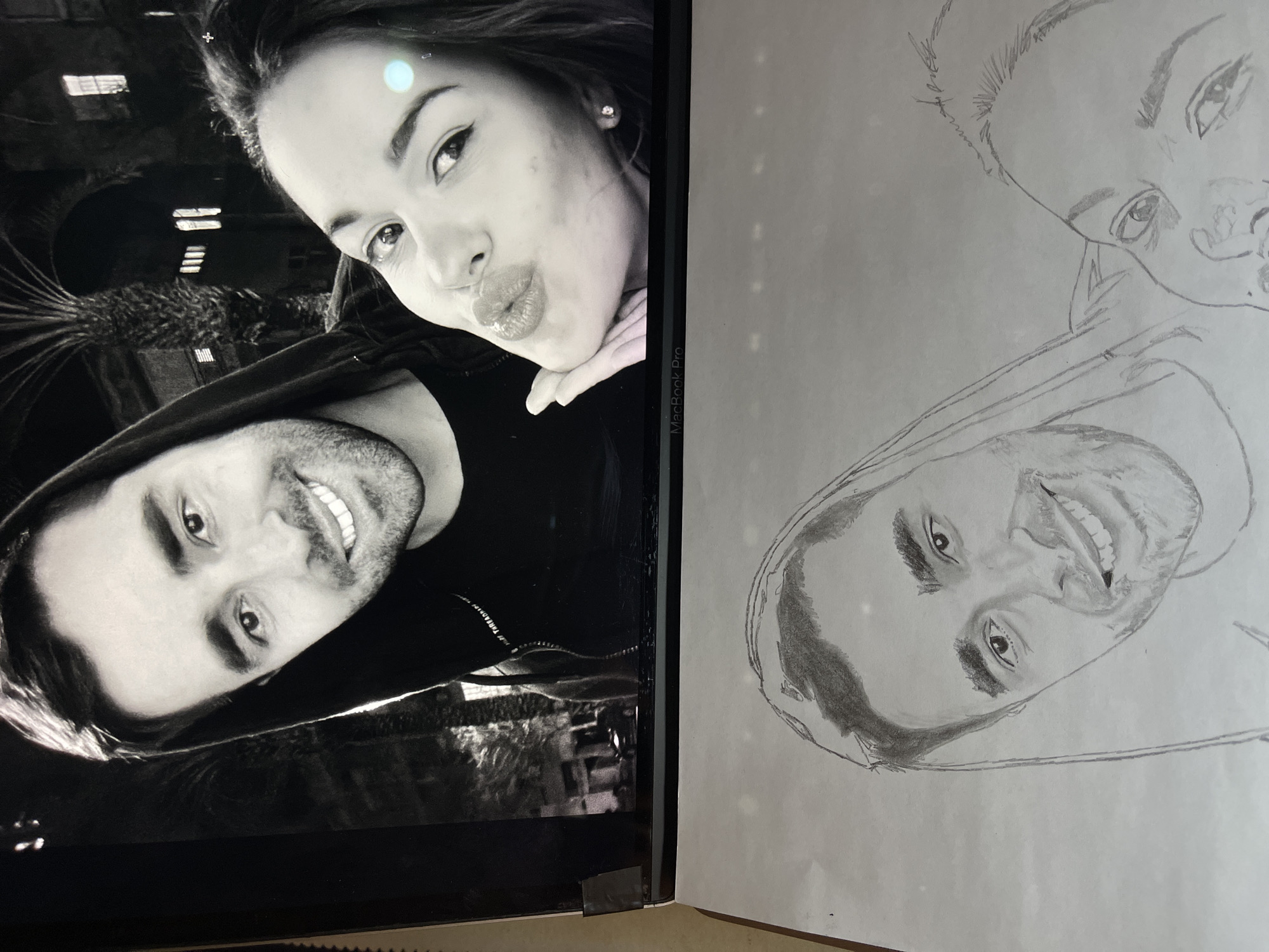 A digital screen, identified as a "MacBook Pro," displays a black and white photograph positioned above a graphite pencil sketch of the same image, resting on a flat surface.
The black and white photograph on the screen depicts two individuals in a close-up composition. On the left, a man with short dark hair, a light beard, and a wide smile looks towards the viewer. He wears a dark, hooded garment. On the right, a woman with dark hair and a light complexion poses with puckered lips, as if blowing a kiss, her left hand positioned near her chin. A small, round earring is visible in her right ear. The background of the photograph is dark, revealing silhouetted palm trees and distant illuminated windows within buildings, suggesting a nighttime urban or semi-urban setting.
Below the screen, a graphite pencil sketch on white paper replicates the photograph. The sketch shows the smiling man and the woman with puckered lips, mirroring their poses from the digital image. The sketch appears to be a work in progress, exhibiting varying levels of detail.
The scene provides no discernible landmarks, specific architectural features, or other identifying elements that confirm a location within the city of Jos, Nigeria.