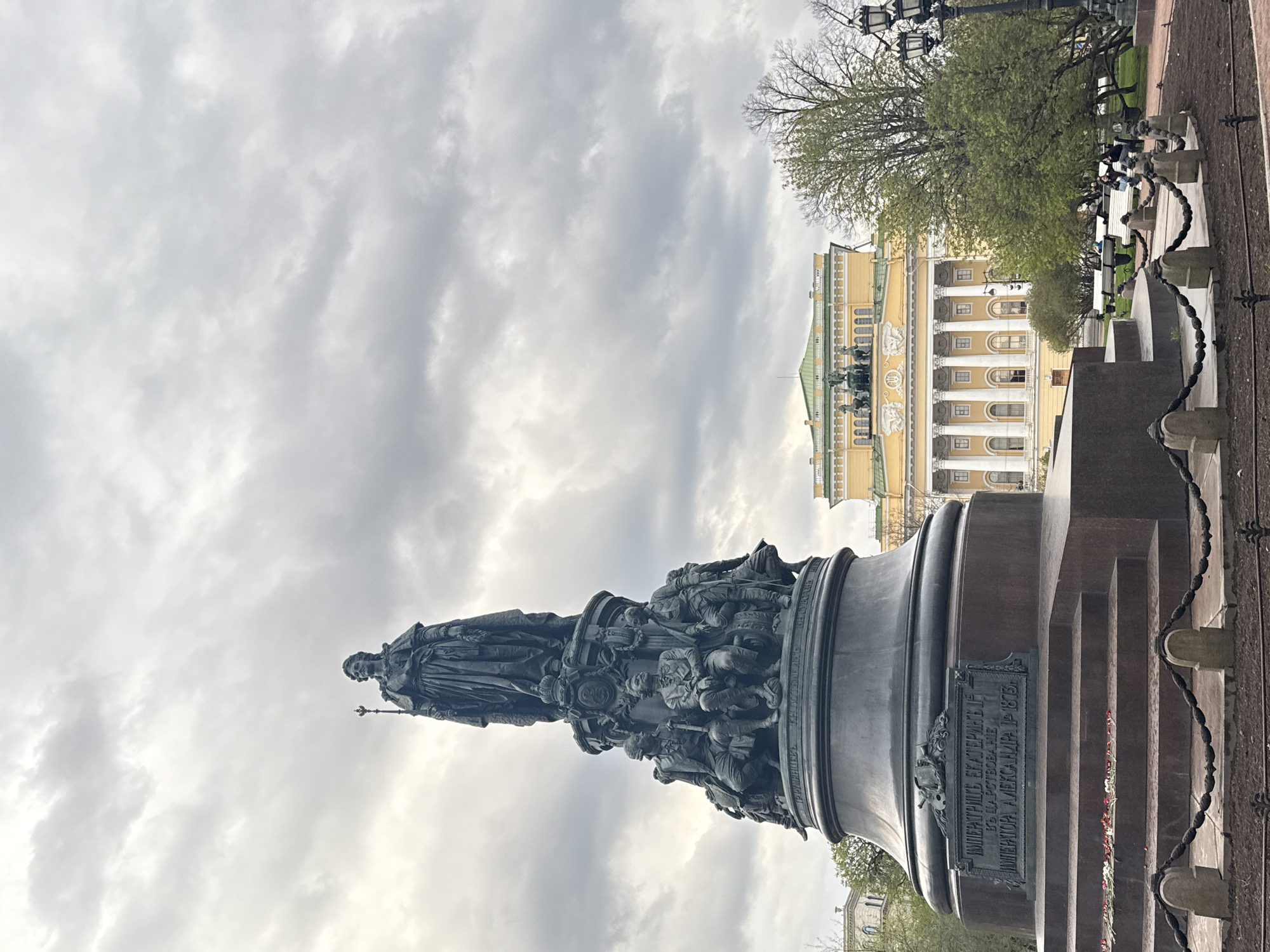 The image depicts the Catherine the Great Monument in St. Petersburg, Russia, with the Hermitage Museum in the background. The monument features a bronze statue of Catherine the Great standing atop a pedestal adorned with figures. The sky is cloudy. To the right are trees and street lamps. People are sitting on benches nearby. The pedestal has a plaque with Russian text. A metal chain fence surrounds the monument.
