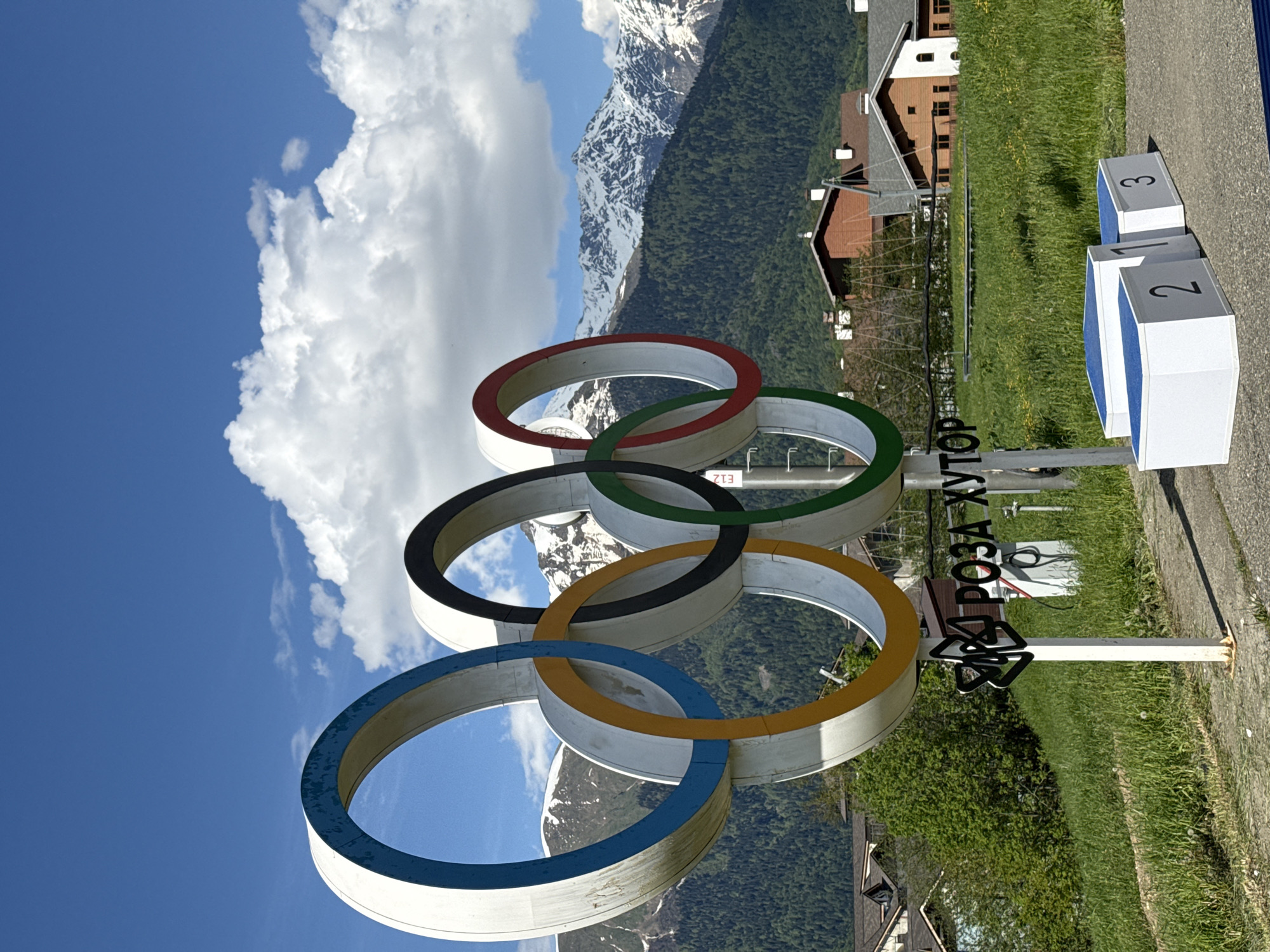 This image captures a vibrant, sunny outdoor scene in Estosadok, Russia, focusing on prominent Olympic landmarks against a dramatic mountain backdrop.
In the foreground, a large, white sculpture of the five Olympic rings stands prominently. The rings are colored red (top left), black (top center), green (top right), yellow (bottom center), and blue (bottom left), intertwined in their traditional arrangement. To the right of the rings, a white sign with black Cyrillic lettering reads "РОЗА ХУТОР" (ROZA KHUTOR), indicating the location as the Rosa Khutor alpine resort, a key venue for the 2014 Sochi Winter Olympics. Further to the right, a winner's podium made of white blocks with blue tops displays the numbers "1", "2", and "3", signifying the first, second, and third place.
The setting is a picturesque mountainous region. In the mid-ground and background, majestic snow-capped mountains rise under a clear, bright blue sky dotted with large, white cumulus clouds. The lower slopes of the mountains are covered in dense green forests, contrasting with the white peaks. Several chalet-style buildings with brown roofs are visible among the trees and on grassy areas, suggesting a resort village. The immediate foreground features lush green grass.
The image portrays a bright day, likely late morning or early afternoon, with strong sunlight casting clear illumination on the scene. No people are present, allowing the focus to remain on the impressive natural landscape and the commemorative Olympic structures. The overall atmosphere evokes a sense of grandeur and sporting heritage.