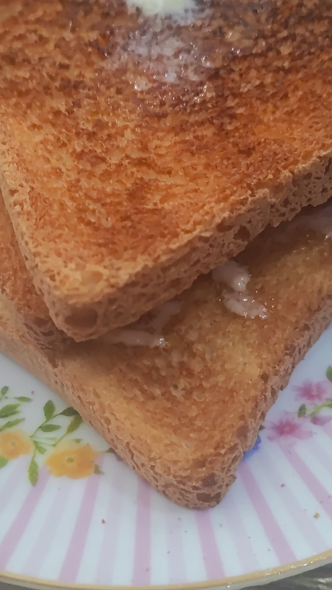 Two slices of toasted bread are stacked on a white and pink striped plate with floral accents. Butter is melted and glistening on the toast. The scene is a close-up, indoor shot, and the background is out of focus. There is no information available about the location in the city of Sochi, Russia.