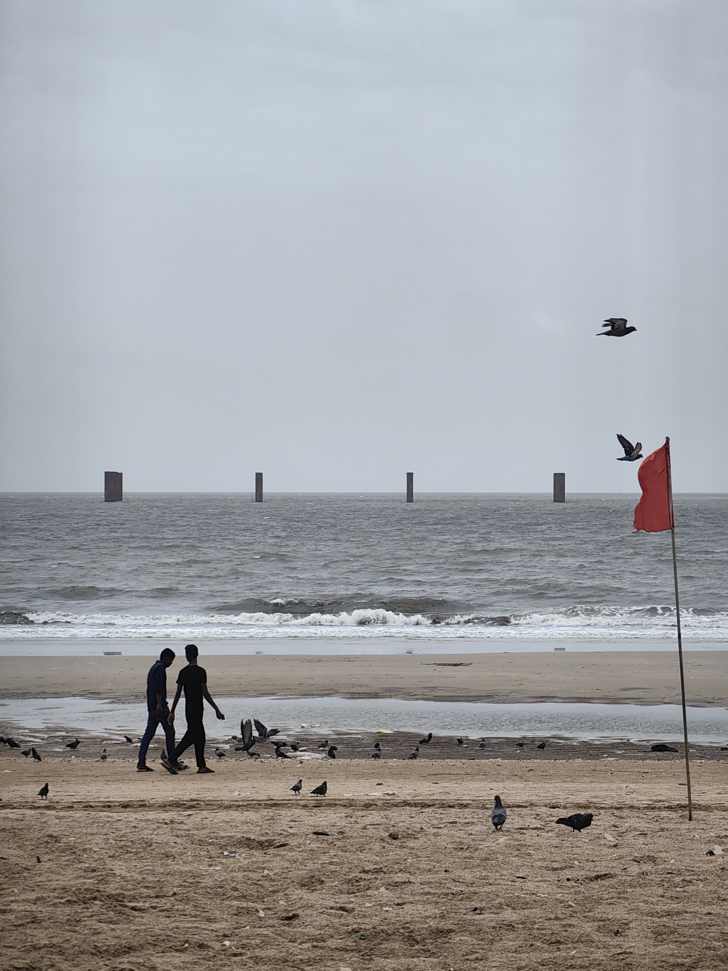 This image depicts a desolate beach on a cloudy day, likely in Mumbai, India, given the presence of similar structures found along the city's coastline. The scene is dominated by a wide, choppy sea under a grey sky. In the distance, four tall, dark, rectangular pillars rise out of the water, spaced evenly across the horizon. The foreground features a sandy beach with scattered debris and several pigeons. Two men are walking from left to right across the mid-ground of the beach, their figures silhouetted against the lighter sand. To the right of the frame, a red flag on a tall pole flutters in the wind. Two birds are in flight in the sky above the sea, with a third bird visible closer to the red flag. The overall atmosphere is somber and somewhat melancholic, with the waves crashing on the shore and the overcast sky contributing to a muted color palette.