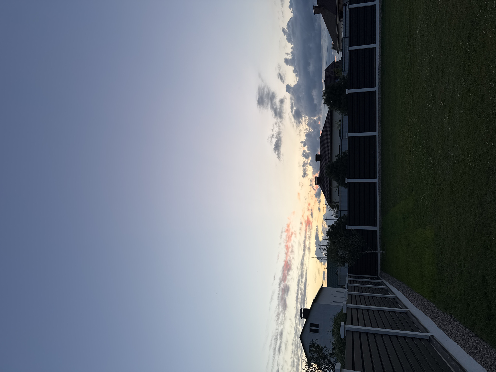 The image shows a suburban residential area with houses and fences under a twilight sky. The scene appears to be located in the Бараўлянскі сельскі Савет region of Belarus. In the foreground, a well-maintained lawn is bordered by a dark horizontal slat fence with white concrete posts. Beyond the fence, several houses with dark tiled roofs are visible, with chimneys. The sky is a gradient of pale blue at the top to pale yellow and pink near the horizon, with scattered dark clouds. The lighting suggests it is either dusk or dawn. There are no people or animals visible in the image. The environment is quiet and appears to be a residential neighborhood.