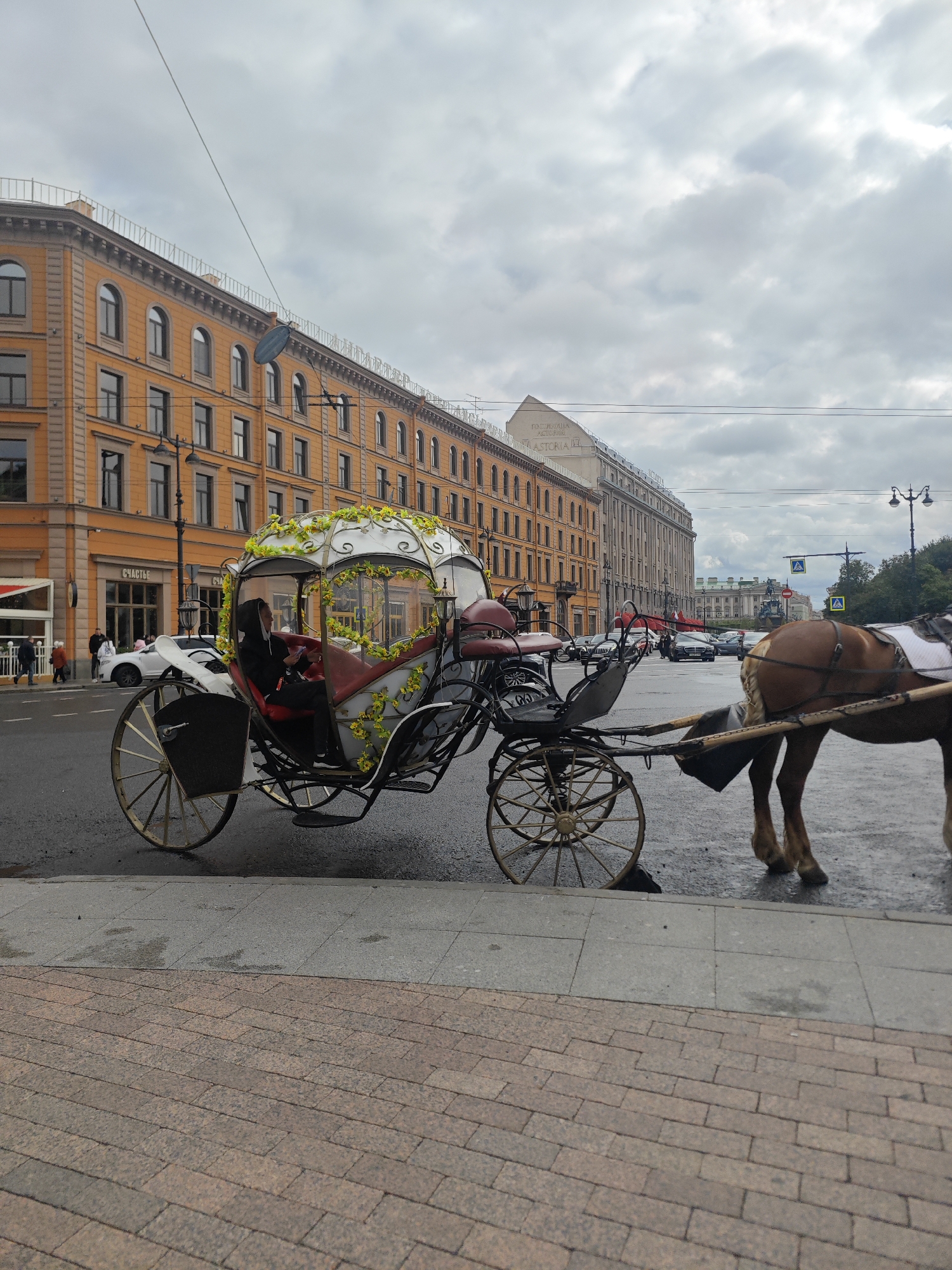 The image depicts a horse-drawn carriage on a city street in Saint Petersburg, Russia. The carriage is ornate, with a white canopy decorated with yellow flowers and intricate metalwork. A person wearing a dark hoodie is seated in the carriage, holding a phone. A brown horse is harnessed to the front of the carriage, visible from the rear.
In the background, there is a long, multi-story building with an orange facade and arched windows. Further down the street, other buildings, traffic lights, and cars are visible, indicating an urban environment. The sky is overcast with clouds. The street appears to be wet, suggesting recent rain. The foreground shows a pedestrian walkway made of brick pavers. The scene is likely a tourist area in Saint Petersburg, given the presence of a horse-drawn carriage and the architectural style of the buildings.
