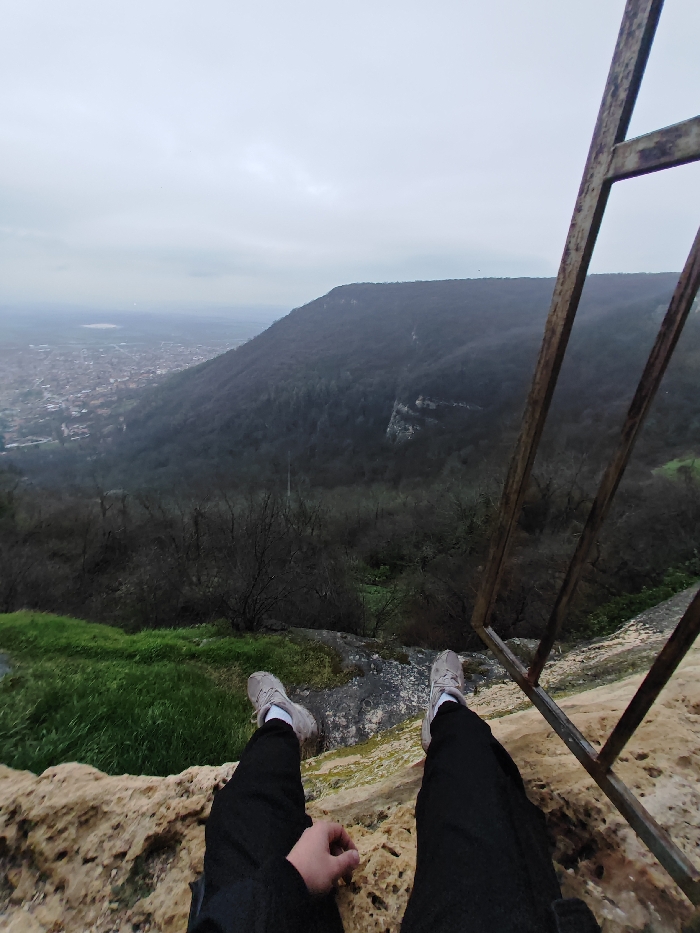 The image presents a first-person perspective from a high vantage point on the Shumen Plateau, overlooking the city of Shumen, Bulgaria. In the immediate foreground, an individual's lower extremities are visible: legs clad in black pants, white socks, and light grey sneakers, extended over a rough, light-colored rocky ledge. The individual's right hand rests on this rocky surface. To the right, a dark, weathered metal railing, possibly part of an observation structure, partially frames the scene. Below the foreground, a steep descent into a valley is covered by a dense, deciduous forest, where trees exhibit minimal foliage, suggesting late autumn, winter, or early spring. Further in the mid-ground and background, the city of Shumen is visible, characterized by a dense arrangement of buildings with reddish-brown roofs. Beyond the city, a flat, hazy plain extends into the distance. The sky is uniformly overcast and grey.