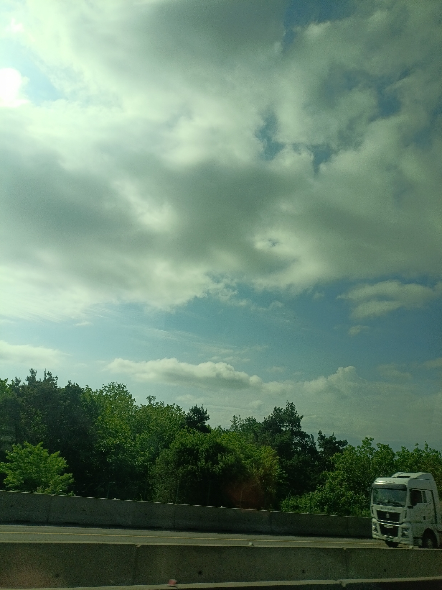 The image shows a roadside scene. A white truck is driving on a multi-lane highway, separated from a forested area by a concrete barrier. The sky is overcast with a mix of white and gray clouds, with patches of blue sky visible. There is no indication of the location being Tlustenkhabl, Russia.