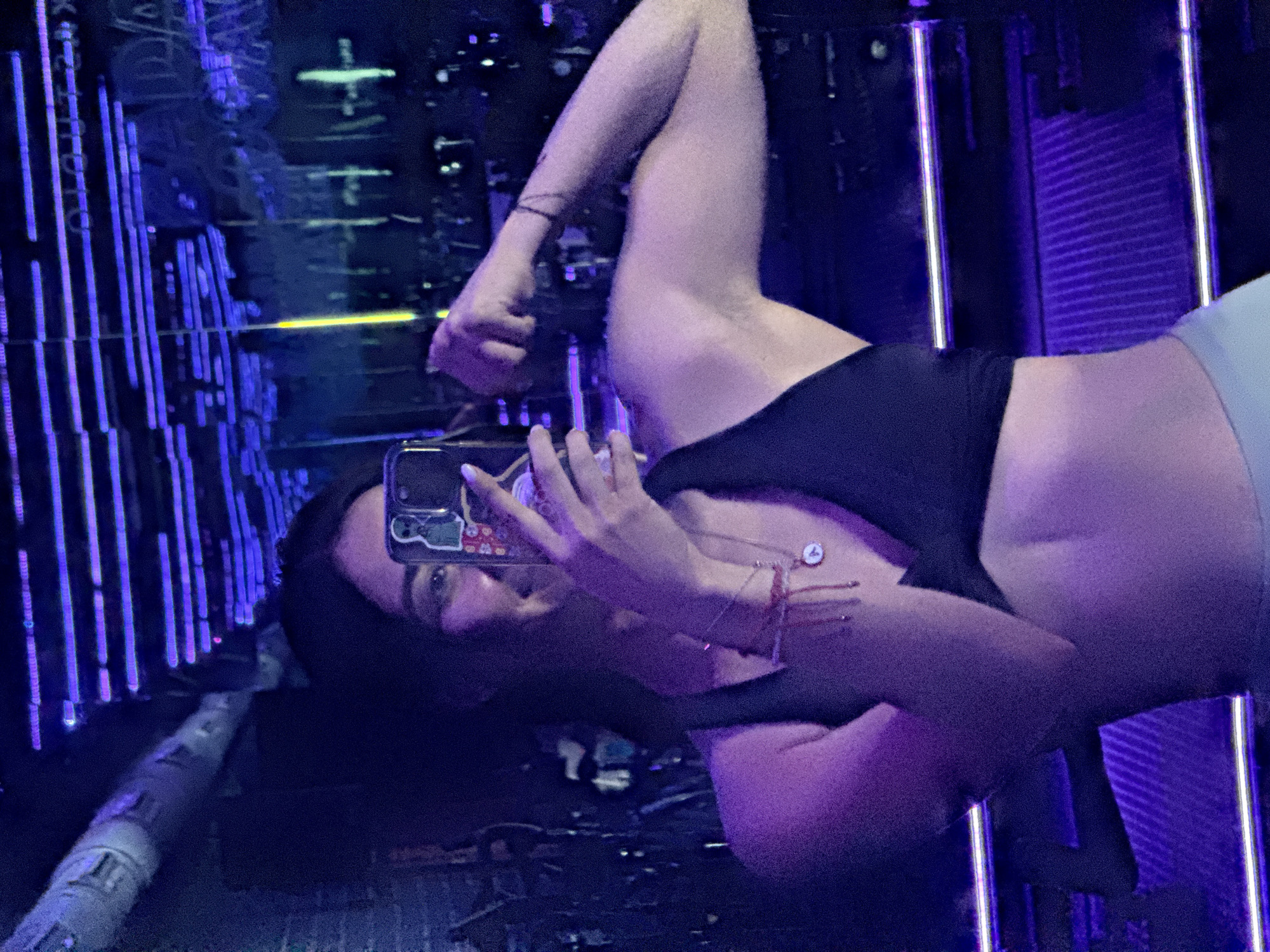 A woman is taking a selfie in a neon-lit environment, possibly in Puebla, Mexico. She is holding a smartphone with stickers on the back and wearing a black top and light-colored bottoms. The background features vertical rows of blue and purple lights, creating a visually striking, futuristic atmosphere. The setting appears to be a public space or installation.