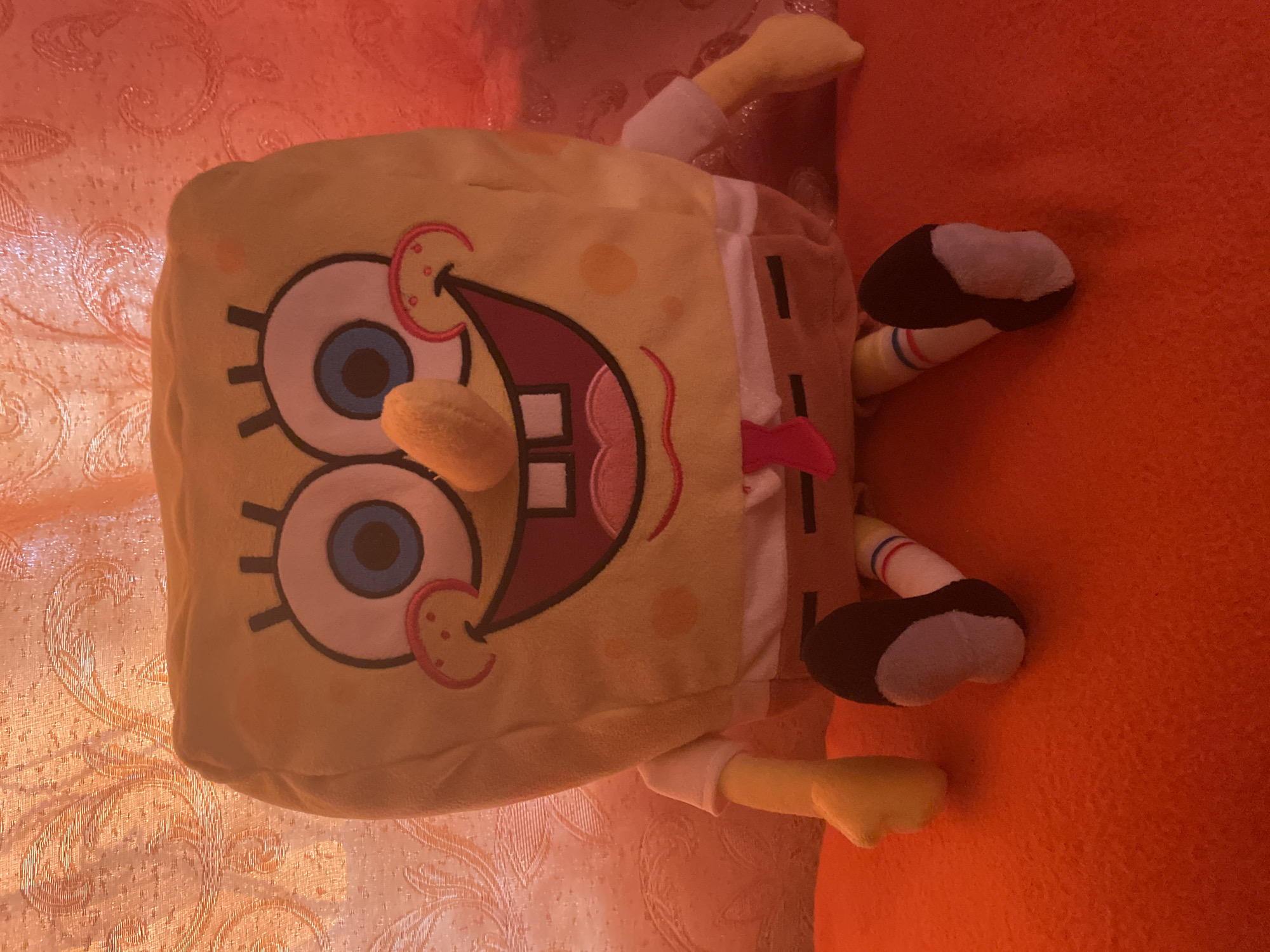 A Spongebob Squarepants plush toy is positioned in a seated posture on a bright orange surface. The toy is yellow with characteristic Spongebob features, including large blue eyes, a wide smile revealing teeth, and rosy cheeks. It is wearing a white shirt, brown pants, and a red tie. Its feet are clad in white socks with blue and red stripes and black shoes. The background consists of an orange textured curtain with an ornate pattern. The scene appears to be indoors. There are no people visible in the image, and no specific location in the city of Shpykolosy, Ukraine can be identified.