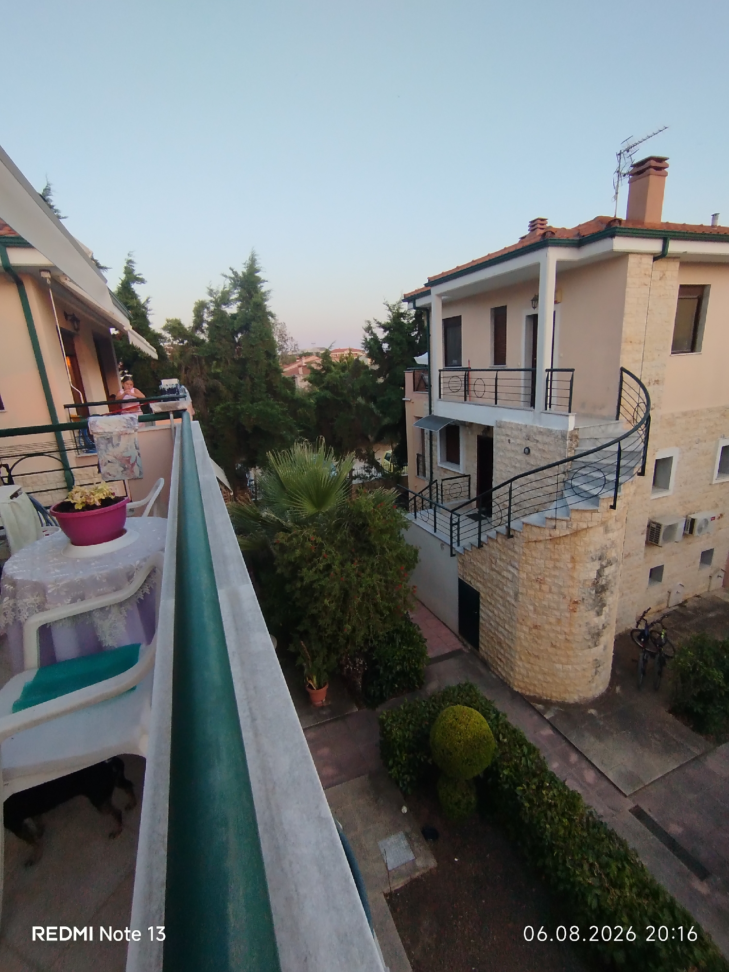The image depicts an outdoor scene, likely a residential building's exterior in Sithonia Municipal Unit, Greece. The foreground features a balcony with a table and chairs, a potted plant, and a dog lying down. To the right, a multi-story building with light-colored walls and a red-tiled roof is visible. The building has several windows, balconies, and an external spiral staircase with a black railing. Air conditioning units are mounted on the building's exterior. In the background, lush green trees and the sky are present. The overall lighting suggests it is either early morning or late afternoon. The timestamp "06.08.2026 20:16" is visible in the lower right corner.
