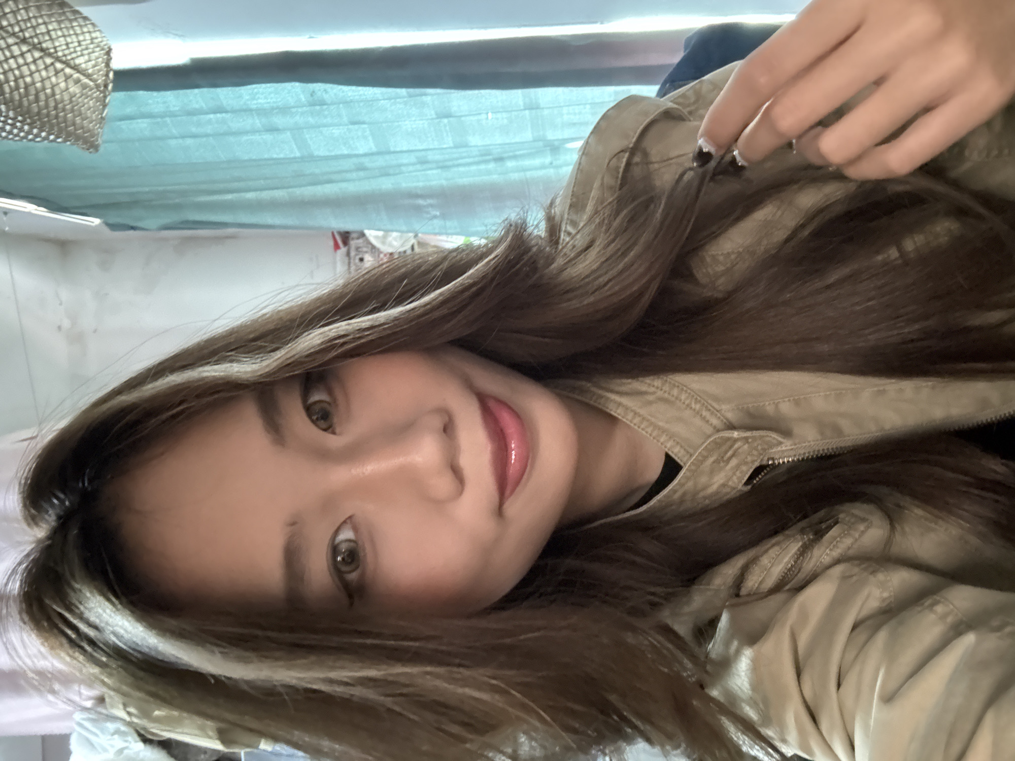 A young woman with long brown hair is posing for a selfie. She is wearing a light brown jacket. Her nails are painted with a black and silver design. The background appears to be an indoor setting with a turquoise curtain. The location in the city of Nan'an District, China, cannot be determined from the image.