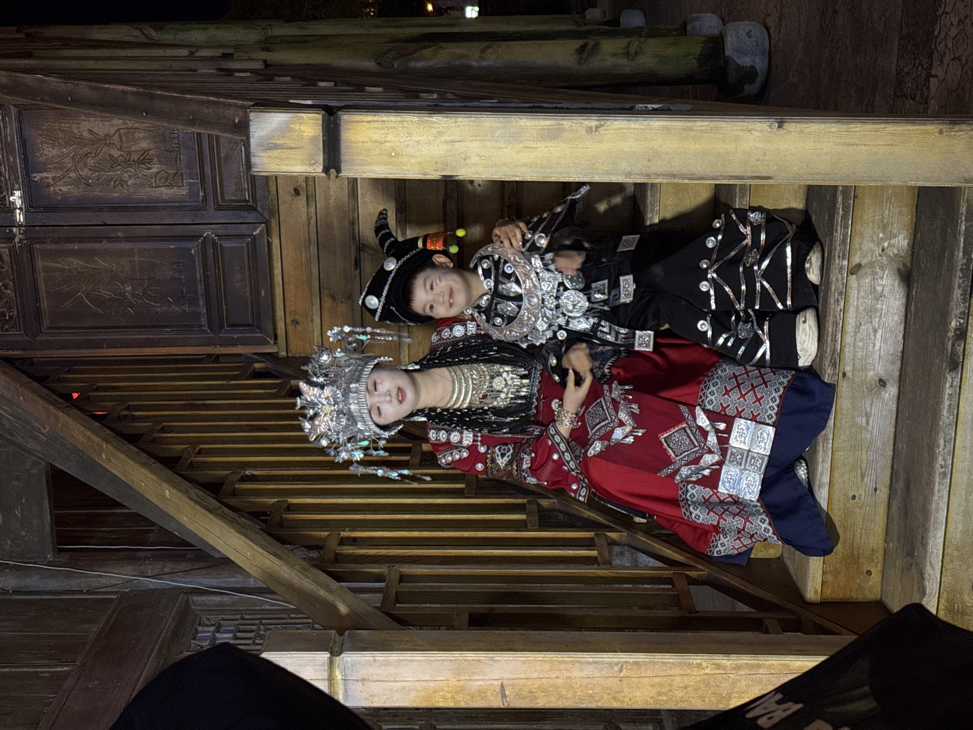 Two individuals are positioned on wooden steps within what appears to be an enclosed, traditional structure. An adult female, adorned in elaborate traditional attire including a large silver headdress, heavy necklaces, and a red patterned robe, is lying on the steps next to a child dressed in a black costume with silver embellishments and a distinctive tall, curved hat. The structure features aged wooden planks and beams, with dark wooden panels on the left. The setting suggests a cultural performance or a staged event taking place in a historic area or building. The individuals are lying, not standing or sitting, on the steps. There is no definitive information in the image to confirm its location in Zhuoshui, China, but the traditional clothing suggests a region with distinct cultural customs.