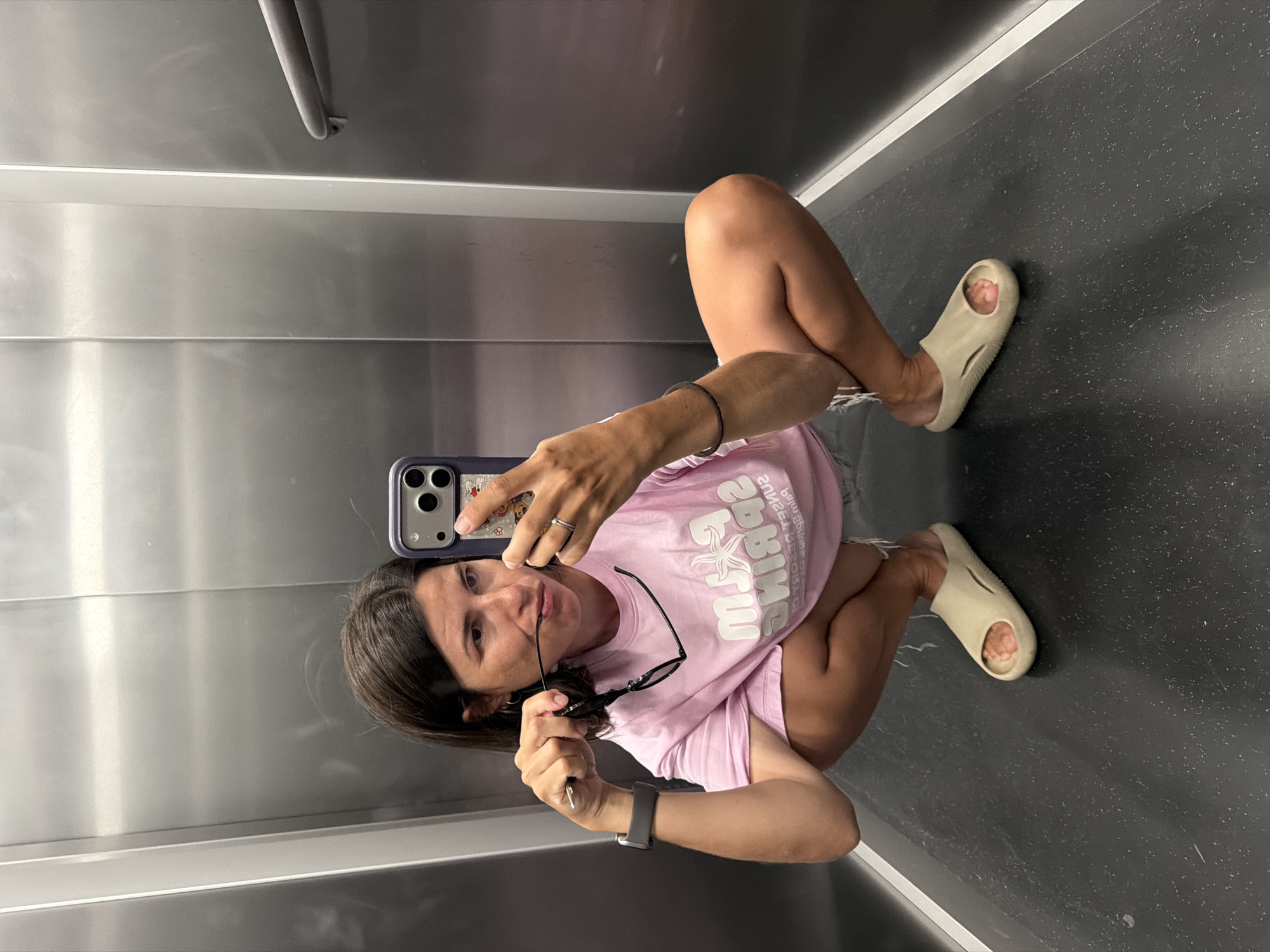 This portrait-oriented image captures a person, likely female, squatting or sitting low on a dark gray, speckled floor within an enclosed space, possibly an elevator or small room. The walls are composed of reflective metallic panels, showing subtle reflections. The subject is looking upwards towards the camera, holding a smartphone with a distinctive case in their right hand, seemingly taking a selfie.
They are dressed casually in a light pink t-shirt with white, mirrored text across the chest. The text is partially legible, appearing to include "ZBEDAS" or "2BEDAS" and other blurred words or symbols. They are also wearing dark shorts with a frayed hem and beige slide sandals. In their left hand, they hold a pair of dark-rimmed sunglasses. Visible accessories include a black watch on their left wrist, a dark elastic band or bracelet on their right wrist, and a silver ring on their right hand. The person has dark hair and a subtle, relaxed expression. The setting is brightly and evenly lit indoors. This casual moment is captured in Budva, Montenegro.