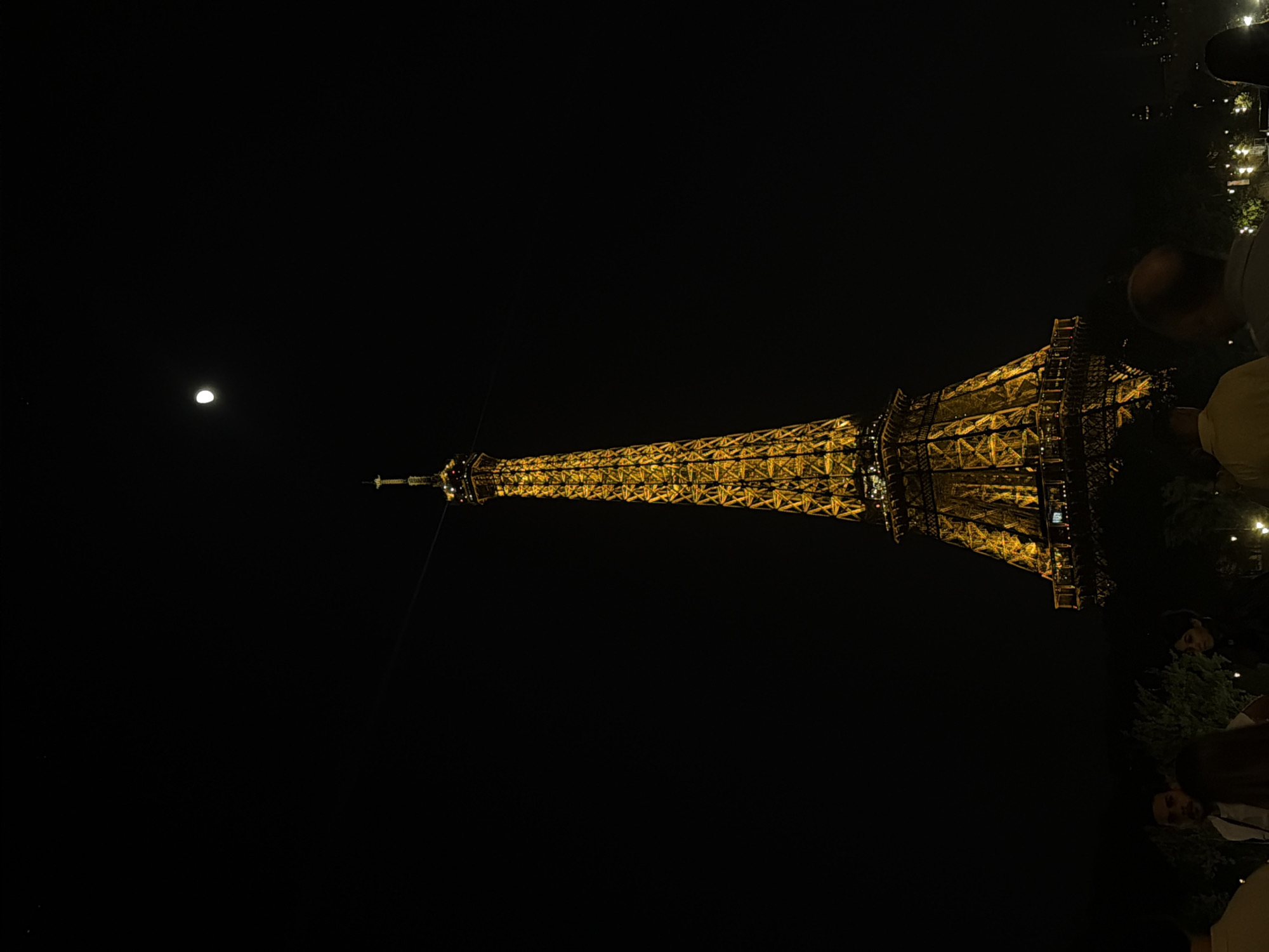 The image shows the Eiffel Tower lit up at night in Paris, France, with a crescent moon visible in the dark sky. Several people are located nearby.