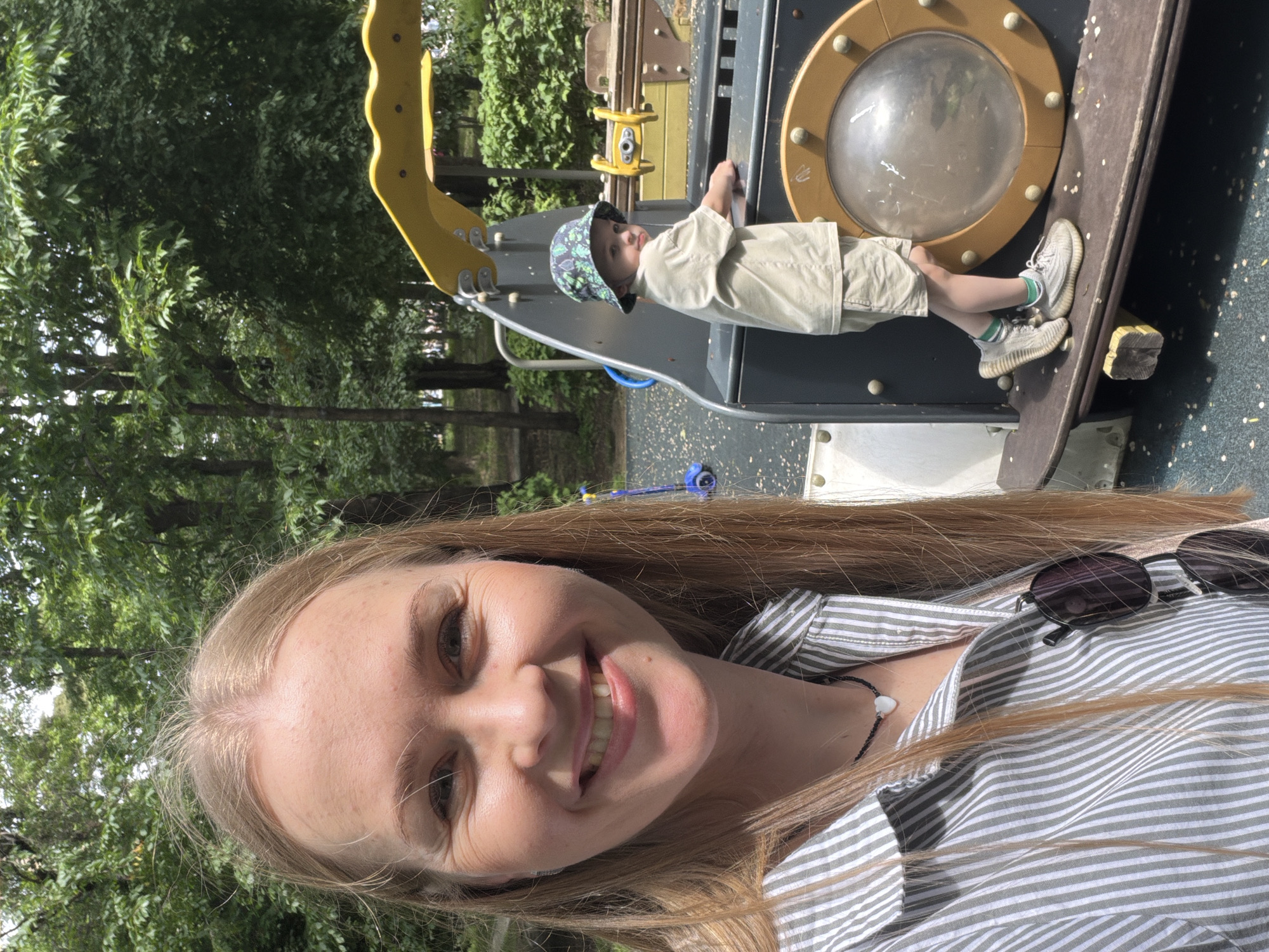 The image shows a young woman taking a selfie outdoors at a playground. She has long blonde hair, is wearing a striped shirt, and is smiling. To her right, a young boy wearing a bucket hat and khaki shorts stands on a playground structure. The playground appears to be located in a park with trees in the background. The overall scene depicts a casual outdoor setting, possibly on a sunny day. The location of Khabarovsk, Russia, is not discernible from the visual information provided in the image.