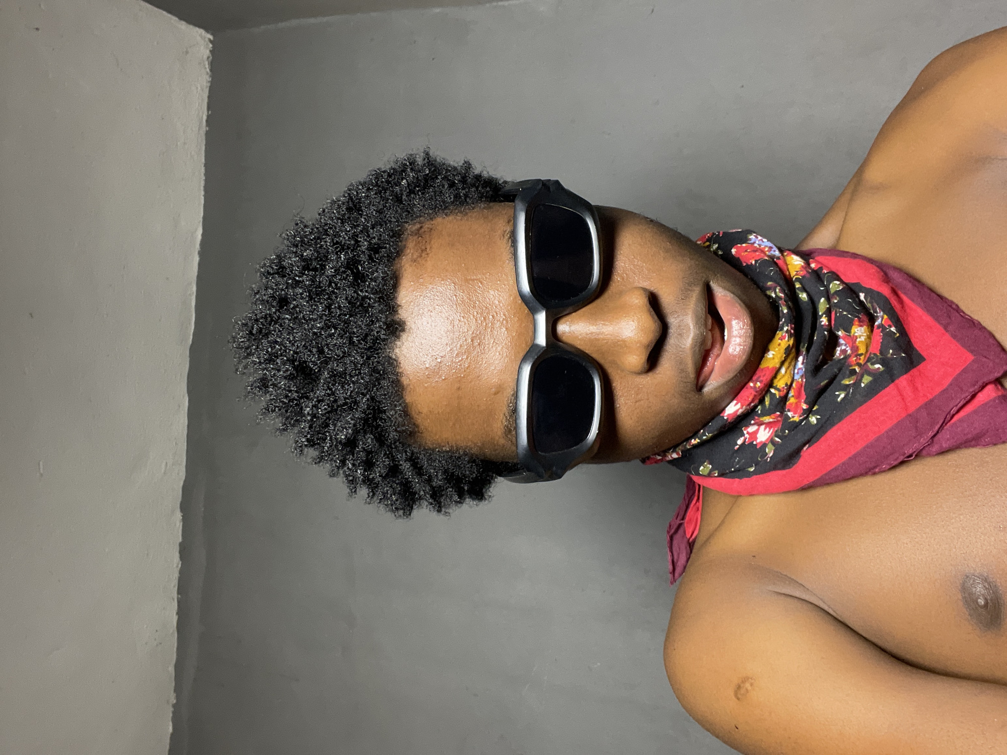 The image shows a close-up, waist-up shot of a young Black male wearing black sunglasses and a colorful bandana around his neck. He has short, black, curly hair and is shirtless. The background is a plain, textured gray wall. The man is looking directly at the camera with his mouth slightly open, and his tongue is visible, appearing to be flicking his lips. His left shoulder and upper chest are visible. There are no other people or objects in the frame. The scene is indoors, likely in a room in Nkpor, Nigeria, given the context provided. The lighting appears to be from above and slightly in front of the subject.