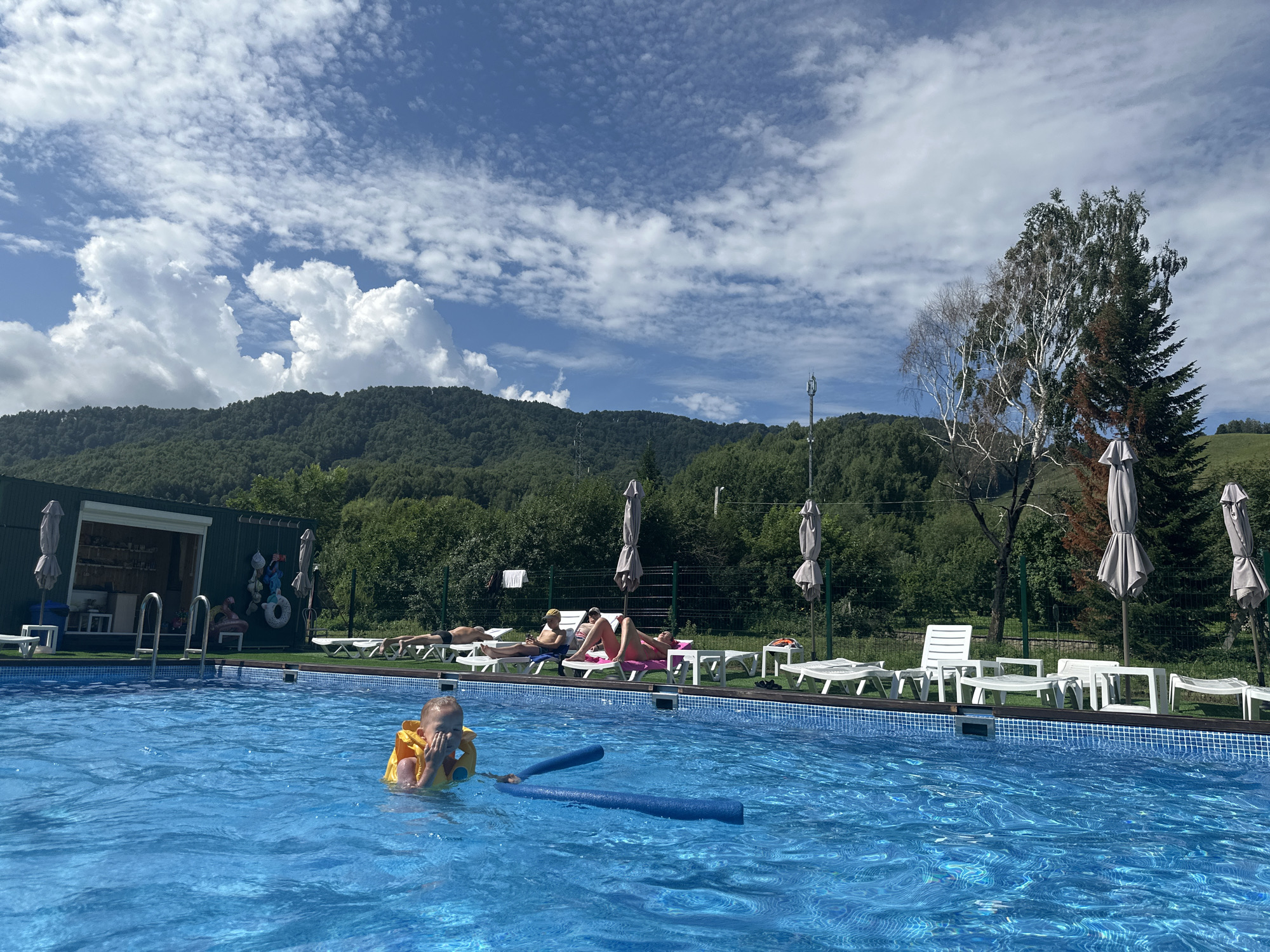 In the city of Danilovka (Altai), Russia, a bright outdoor swimming pool is depicted on a sunny day with scattered clouds. The water is a vibrant blue, reflecting the sky and sunlight. In the foreground, a young child wearing a yellow flotation vest is in the pool, holding blue pool noodles. Several adults are relaxing on lounge chairs by the poolside. Behind the pool, a building resembling a changing room or service area with an open window and various items hanging on the wall is visible, along with several closed umbrellas. The background is dominated by a lush green forested hillside, with a clear blue sky and white clouds above.