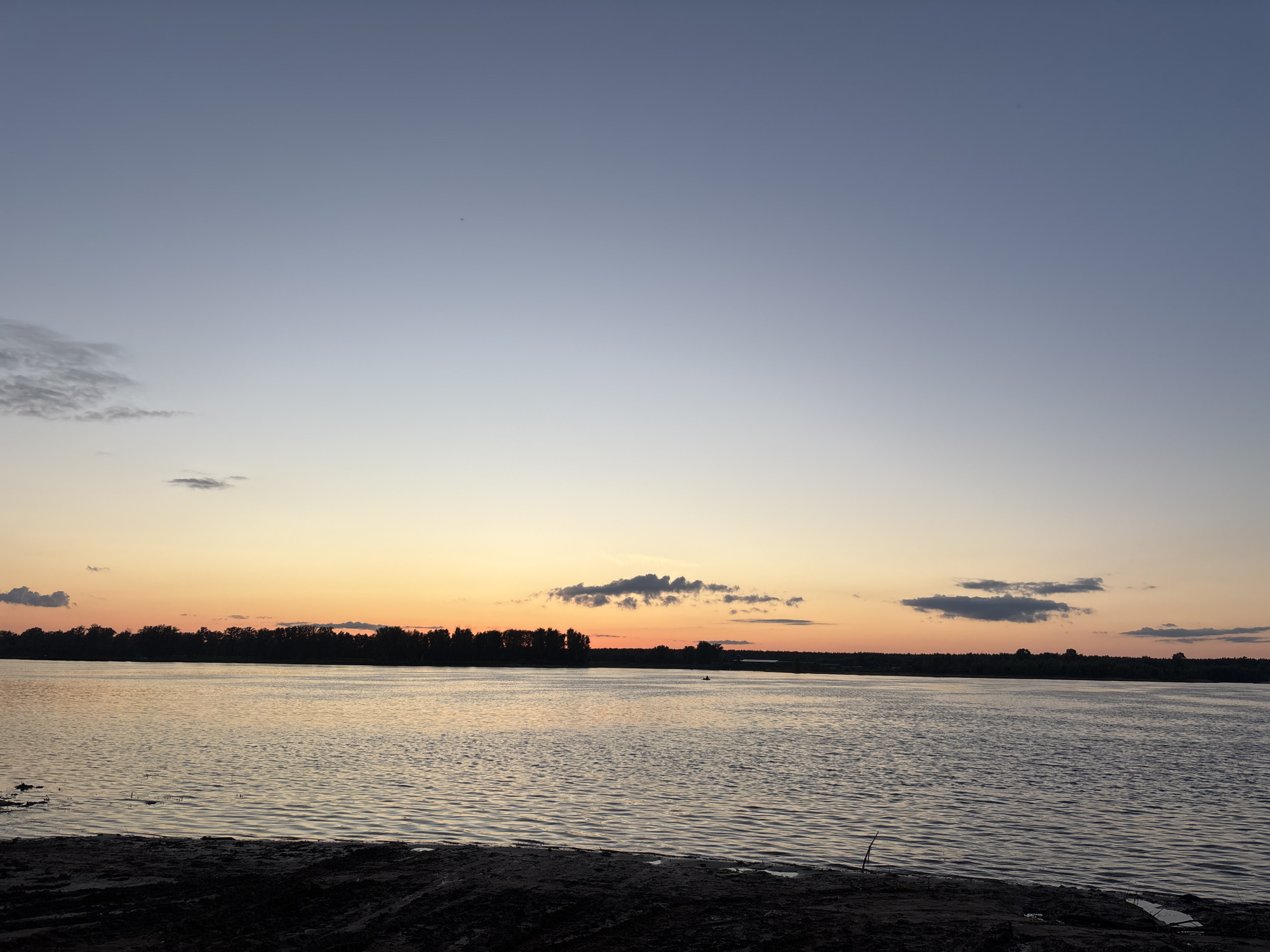 The image depicts a serene sunset over a wide body of water, likely a river, in Olkhovka, Russia. The sky transitions from a soft blue at the top to a warm gradient of orange and yellow near the horizon, indicating the sun is setting. Scattered clouds are present in the sky, some tinged with the colors of the sunset. A dark, silhouetted treeline forms the distant shore, suggesting a natural landscape. The water's surface is rippled, reflecting the colors of the sky. In the foreground, dark, sandy terrain is visible, with some sparse vegetation. A small boat with a single occupant can be seen on the water in the middle distance. The overall impression is one of tranquility and natural beauty.