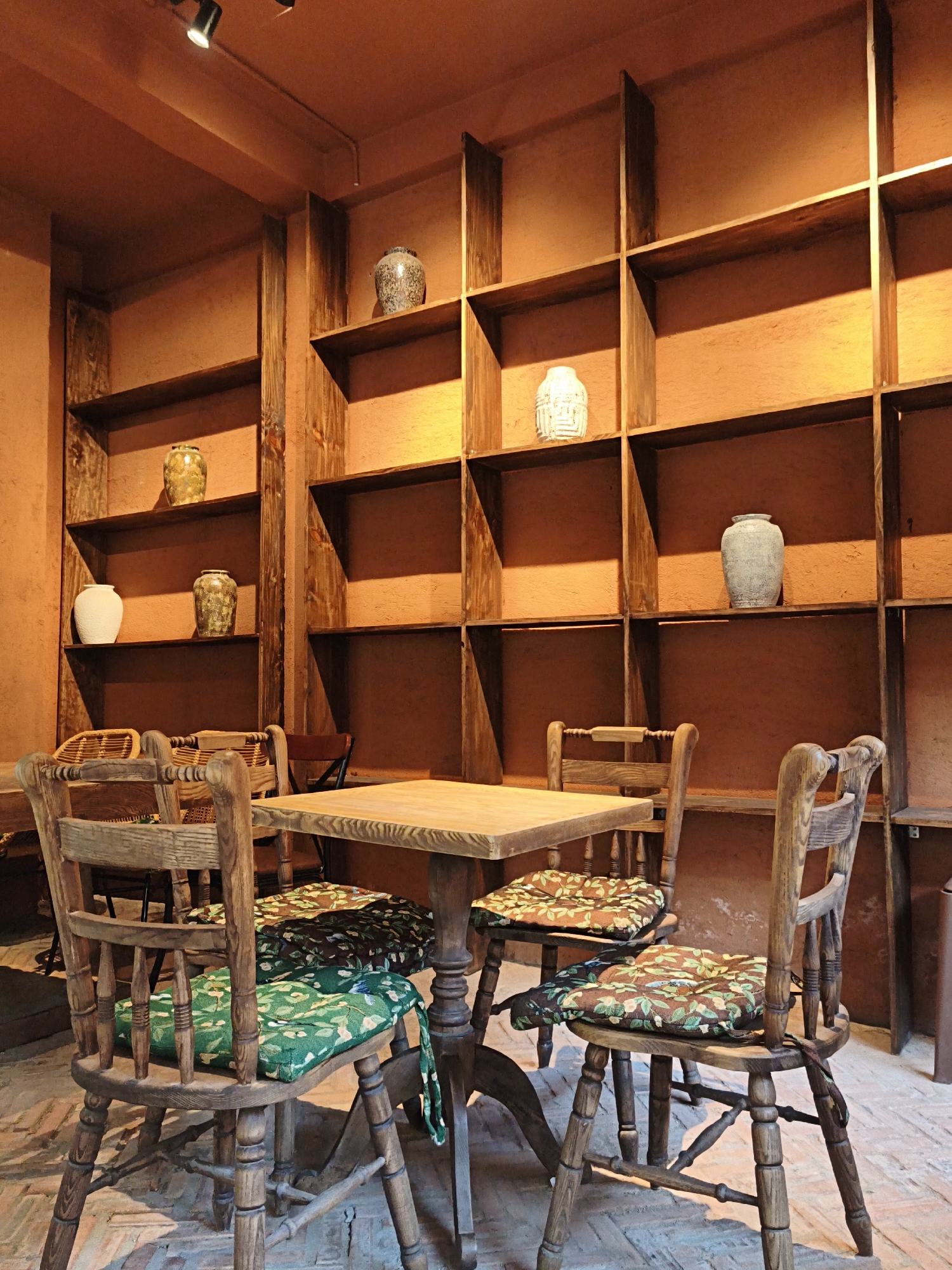 The image depicts the interior of a rustic cafe or restaurant. The dominant feature is a large, multi-section wooden shelving unit that occupies a significant portion of the background. The shelves are filled with various decorative ceramic vases of different shapes, sizes, and colors, including a patterned speckled vase, a white patterned vase, a yellowish vase with green speckles, and a light grey vase. The walls and shelves are painted or finished in a warm, earthy tone, possibly terracotta.
In the foreground, a wooden table with a light-colored surface is set with several wooden chairs. The chairs have a classic, somewhat aged design with turned legs and spindles. Each chair is fitted with a cushioned seat cover featuring a floral or botanical print in shades of green and brown. Two chairs are clearly visible in the front, one with a green patterned cushion and another with a brown patterned cushion. The flooring appears to be brick or a similar textured material.
Overhead, a track lighting system is visible, with two spotlights illuminating the space. The overall ambiance is cozy and natural, suggesting a place with a focus on traditional decor and a relaxed atmosphere. The image does not contain any people, indicating the scene is captured at a time when the establishment is not occupied. The specific location in Bac Nha Trang Ward, Vietnam, is not visually identifiable from the image itself, but the description focuses on the visible elements within the frame.