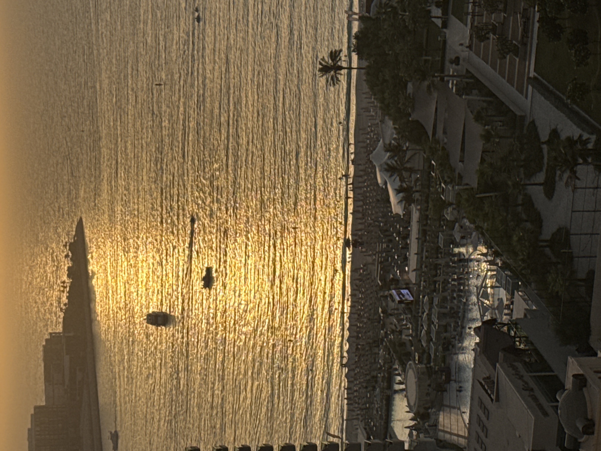 The image captures a coastal view in Dubai, United Arab Emirates, during sunset. The sun's golden light reflects vibrantly on the rippling surface of the ocean. Several boats are visible on the water, moving away from the shore. Along the coastline, a resort area is discernible, featuring a beach lined with rows of umbrellas and lounge chairs, and a swimming pool area where a few people can be seen. Beyond the beach, a lush green landscape dotted with palm trees and various structures, likely part of the resort, extends inland. In the background, a cluster of buildings forming a silhouette against the sunset sky suggests a developed urban or resort complex. The overall scene depicts a tranquil evening at a popular tourist destination.