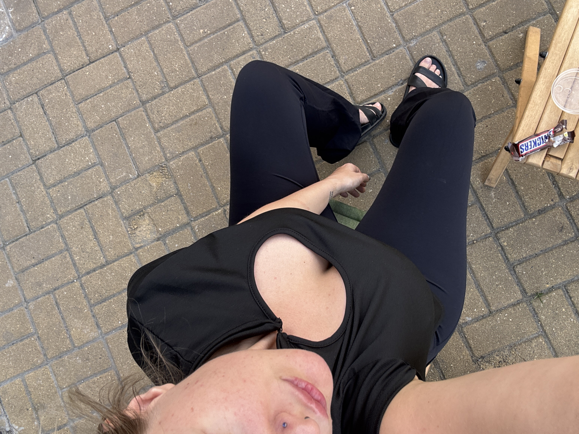 A person, viewed from above, sits on a wooden bench outdoors on a paved patio area with a herringbone brick pattern. The person is wearing a black crop top with a heart-shaped cutout and black flared pants. Black sandals are worn on the feet. On the bench next to the person are two Snickers candy bars and a clear plastic cup. The scene appears to be in an urban outdoor setting. There is no specific indication of the location being Moscow, Russia.