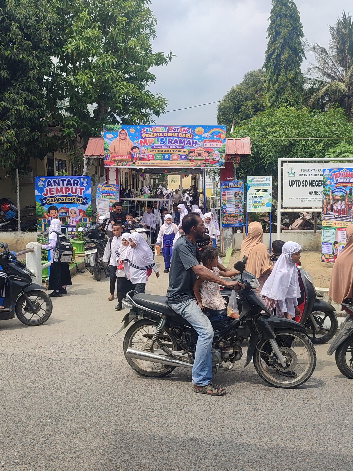 The image depicts an outdoor scene at what appears to be the entrance of a school, likely UPTD SD NEGERI 4 PEUSANGAN in Panton Rah, Indonesia, given the signage. A crowd of people, primarily children in school uniforms, are gathered on a paved road. Many of the children are wearing white shirts, black skirts or trousers, and hijabs, suggesting a religious or cultural context.
Several motorcycles are parked or being ridden, with some individuals on them. One prominent motorcycle in the foreground carries a man wearing a dark t-shirt and blue jeans, with a young girl in a pink and white patterned top on his lap. Another motorcycle is ridden by a man in a black t-shirt and blue jeans. Children are seen walking and standing around, some carrying backpacks.
Large banners are displayed, including one that reads "SELAMAT DATANG PESERTA DIDIK BARU MPLS RAMAH" and another with "ANTAR JEMPUT SAMPAI DISINI." These banners indicate a welcome event for new students, likely related to the "Masa Pengenalan Lingkungan Sekolah" (MPLS), or school environment introduction period. The overall atmosphere appears to be busy and celebratory, with the school day or an event just beginning or concluding.
The environment includes a building structure with a red roof visible behind some of the banners, and lush green trees and palm trees line the background, suggesting a tropical or subtropical climate. The sky is partly cloudy. The road surface is asphalt with visible aggregate. The time of day appears to be daytime, with moderate sunlight casting shadows.