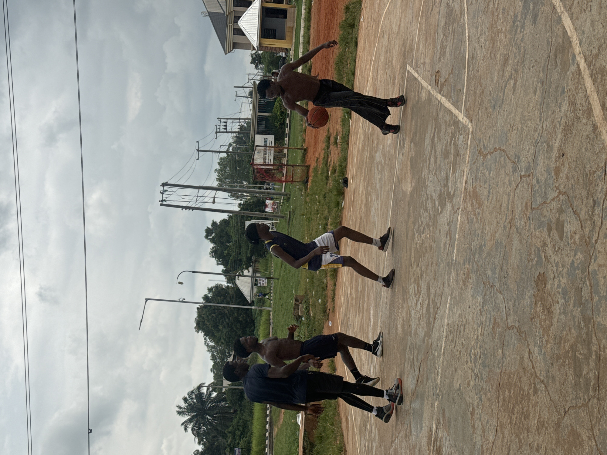 Four young men are playing basketball on an outdoor court in Amansee, Nigeria. The court is made of weathered concrete with white lines marking the boundaries. The sky is overcast, with scattered clouds. In the background, there are trees, utility poles with power lines, and a building that appears to be part of a school or institution, identified by a sign that reads "SCHOOL OF GENERAL STUDIES". One man in the foreground is dribbling a basketball and wearing loose black pants and sneakers. To his left, another shirtless man is running, wearing black shorts and sneakers. Further left, a third man in a blue t-shirt and black shorts with black compression leggings is walking, also wearing sneakers. The fourth man, to the far left, is partially visible and is wearing a blue and yellow sleeveless jersey and white shorts. The overall environment suggests a casual recreational activity in an outdoor urban or semi-urban setting.