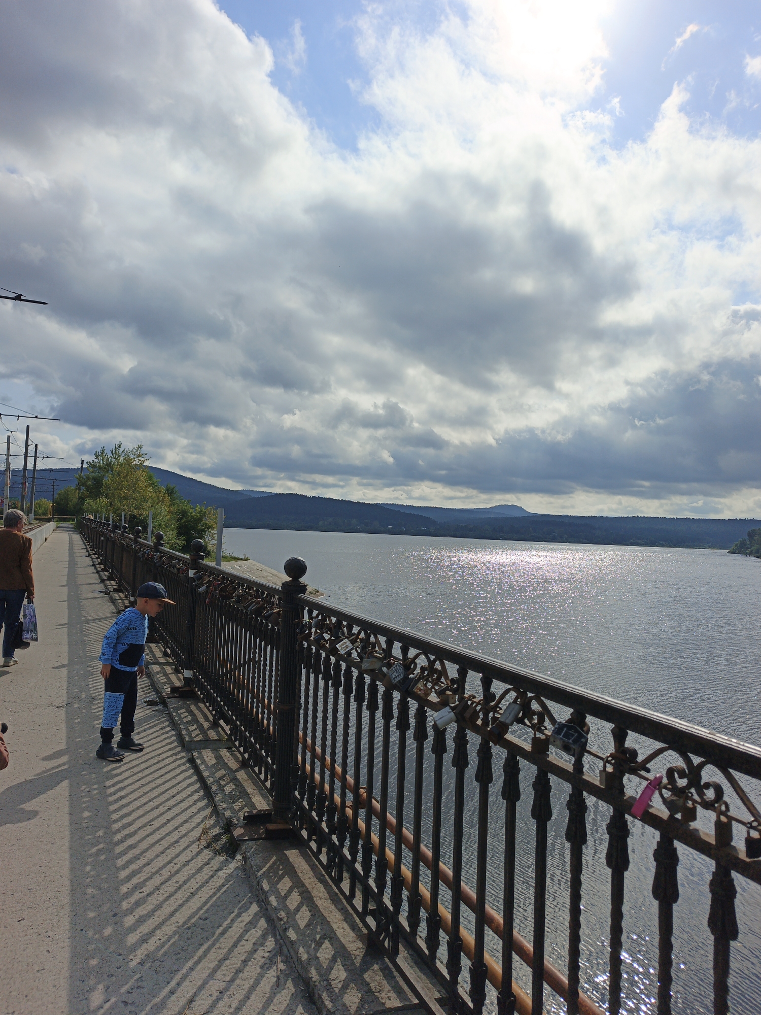 A young boy wearing a blue tracksuit and a baseball cap stands on a pedestrian bridge, looking out over a wide body of water. He is positioned on the left side of the frame, his body angled towards the water. A woman in a brown jacket and blue pants walks away from him further down the bridge. The bridge has a black metal railing adorned with numerous padlocks. The water is shimmering with sunlight, reflecting the bright sky filled with dramatic, grey and white clouds. In the distance, a tree-covered mountain range forms the horizon. The bridge's concrete walkway casts long, striped shadows from the railing onto the ground. Overhead, power lines and poles are visible, suggesting the presence of a tram or trolley system. The scene appears to be a scenic viewpoint in Zlatoust, Russia, likely overlooking a reservoir or large river.