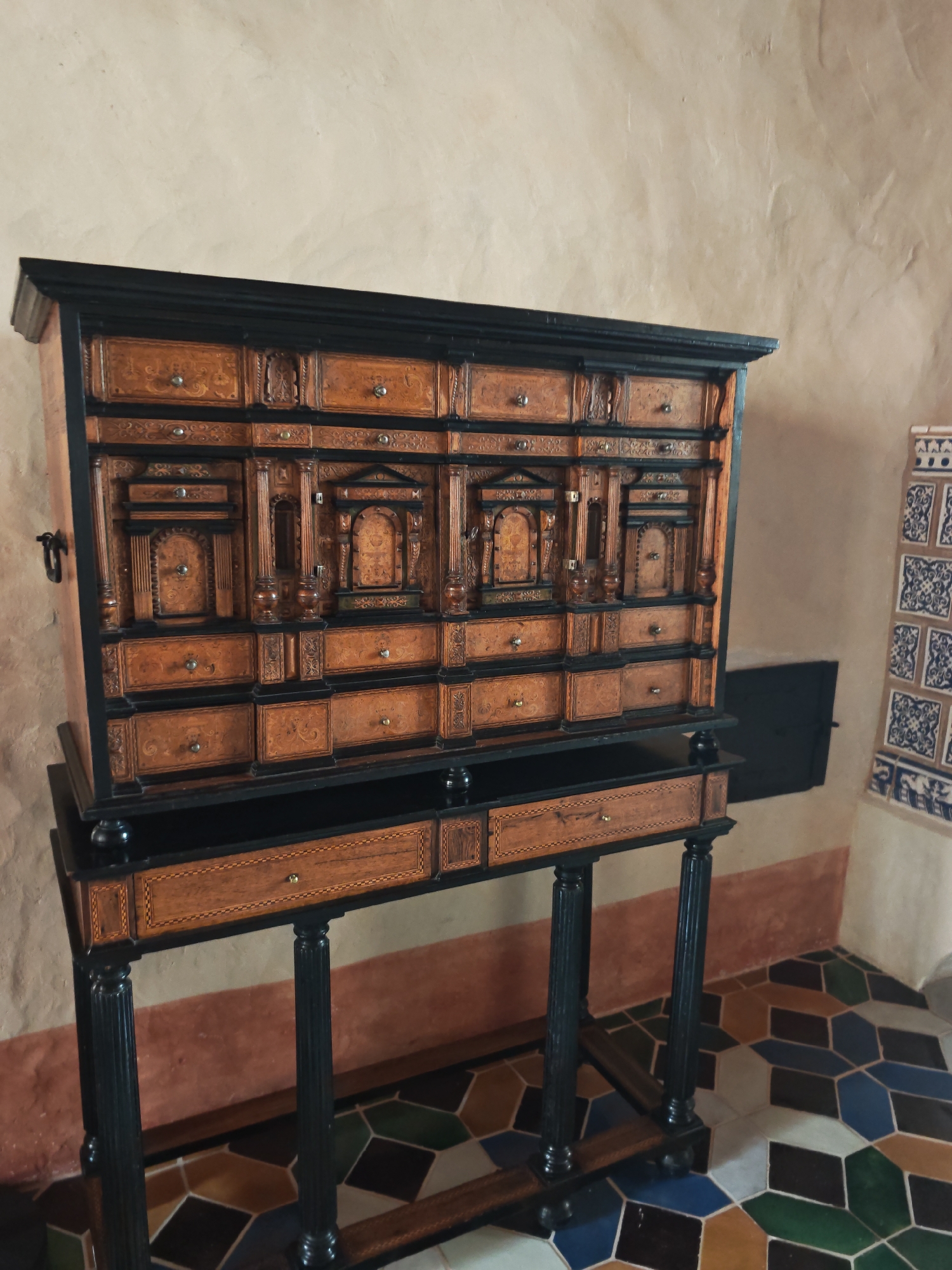 The image shows an antique cabinet with multiple drawers and compartments, set against a textured, light-colored wall. The cabinet is made of dark wood and intricately decorated with lighter wood inlay, featuring geometric patterns and small drawer pulls. It stands on a dark wooden stand with four fluted legs. To the right of the cabinet, a section of tiled wall is visible, adorned with blue and white geometric patterns. The floor is covered in a colorful tessellated tile pattern with hexagonal and rhomboid shapes in shades of brown, green, blue, and white. The scene appears to be indoors, possibly in a museum or historical building. This antique cabinet is on display in the Bauska Castle Museum in Bauska, Latvia, and is a 17th-century Baroque cabinet.