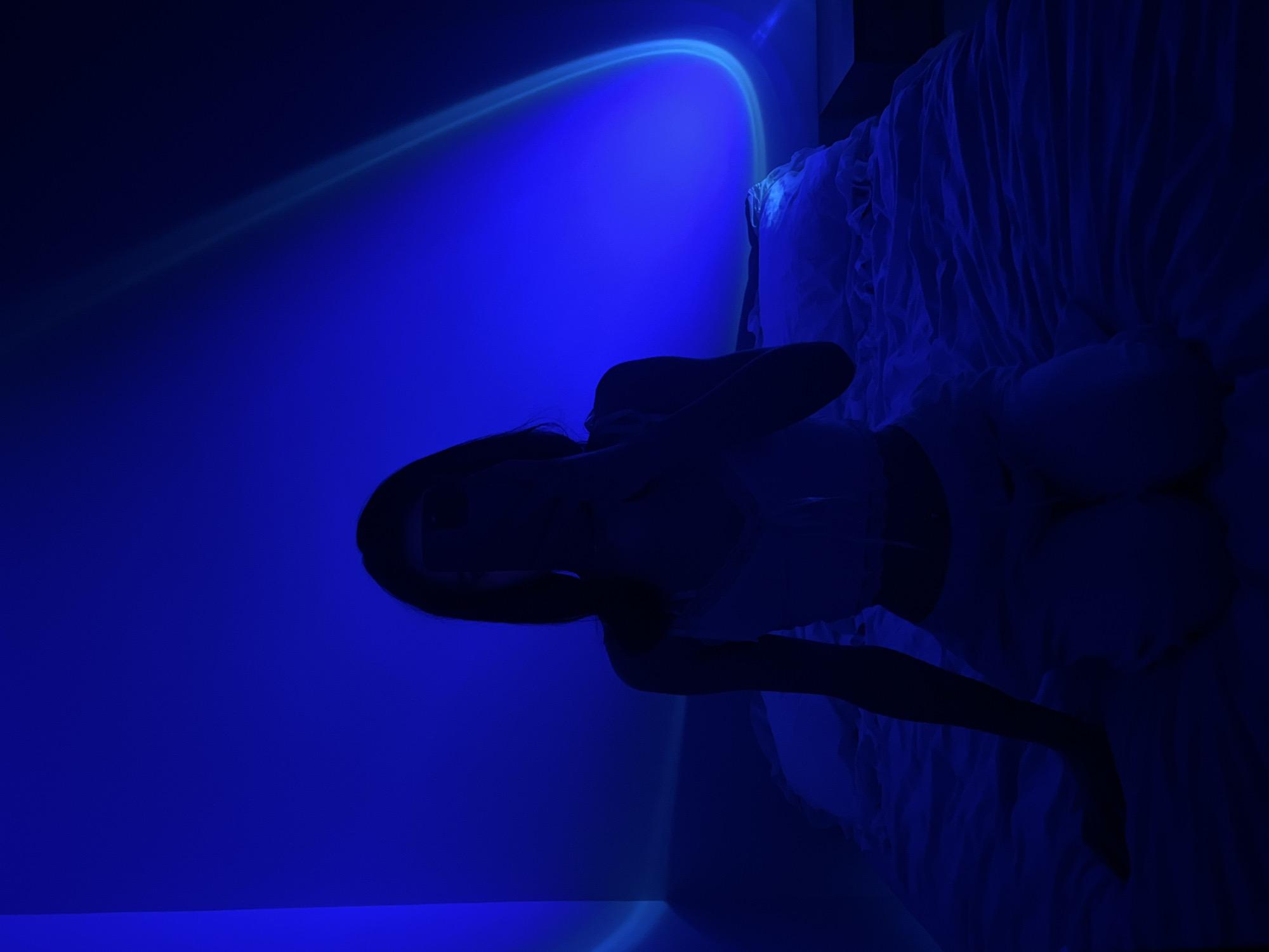 A person is kneeling on a bed with white linens. The room is illuminated by a blue light that casts shadows on the walls and creates a gradient of light and dark. The person is silhouetted against the blue background, wearing what appears to be a white tank top and light-colored pants. Their right hand is raised towards their face, obscuring it. The bed is positioned in the center of the frame, with the person on the left side of the bed, facing the viewer. The background shows a wall with streaks of light. There is no visible indication of the location being Ternopil, Ukraine.
