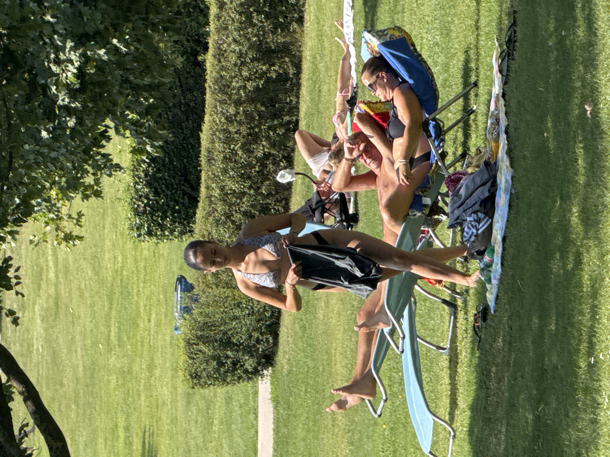 The image captures a sunny outdoor scene at the Hainburg an der Donau leisure center, likely during the summer months. A woman in a bikini stands in the foreground, holding a dark gray towel. She is positioned between two blue lounge chairs, one occupied by a man whose legs and feet are visible, and another empty chair. In the background, several other individuals are sunbathing on lounge chairs and towels scattered across a manicured green lawn. Lush green hedges form a border behind the lawn, and a large tree provides shade in the upper left portion of the image. The atmosphere appears relaxed, with people enjoying the warm weather.