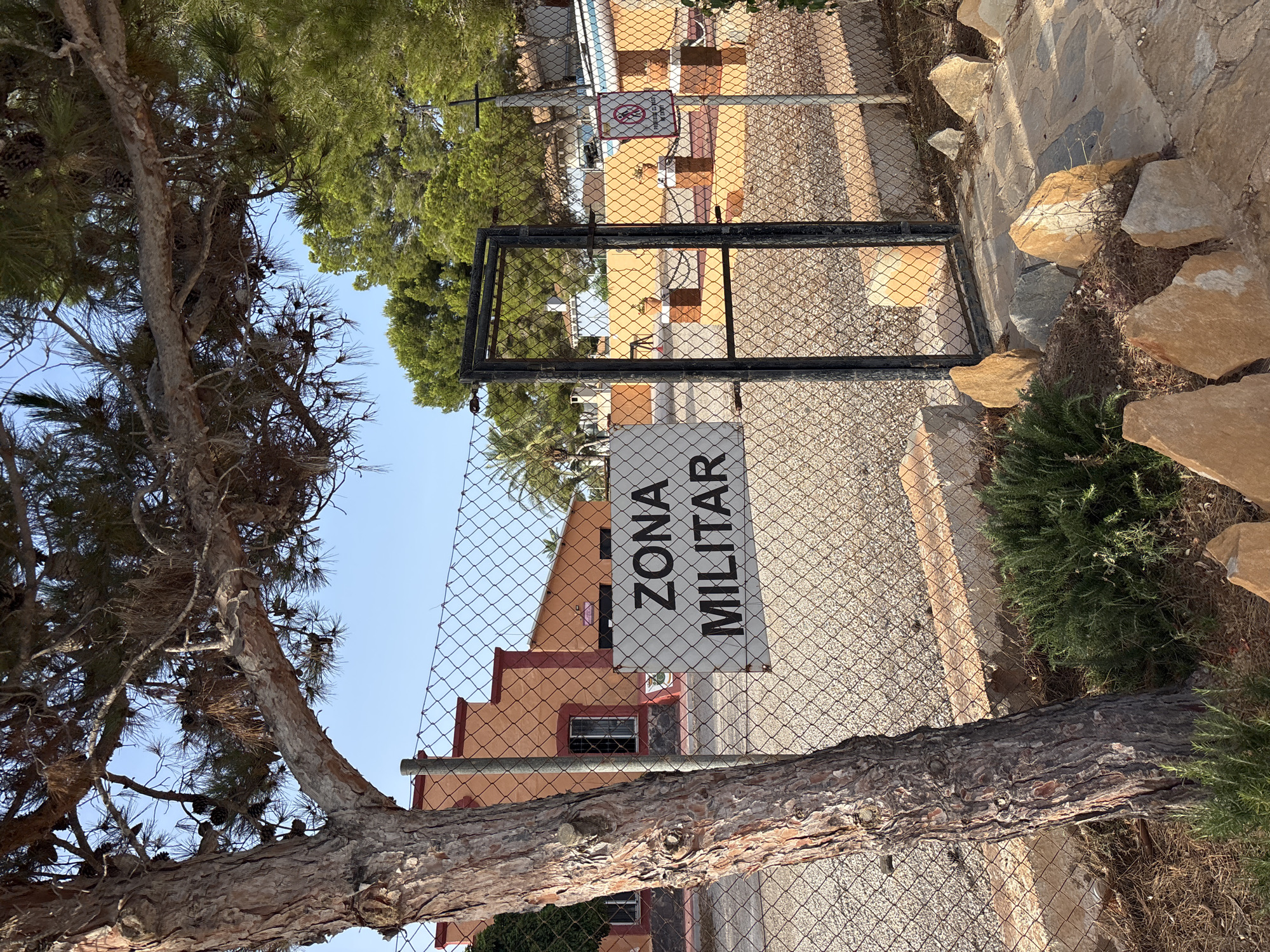 The image depicts a chain-link fence with a gate, behind which is a building and trees. A sign on the fence reads "ZONA MILITAR" in black lettering on a white background. A tree with rough bark stands to the left of the fence, and some rocks and greenery are in the foreground. The building appears to be orange with red trim. The sky is clear and blue. The scene suggests a restricted or protected area within a city.