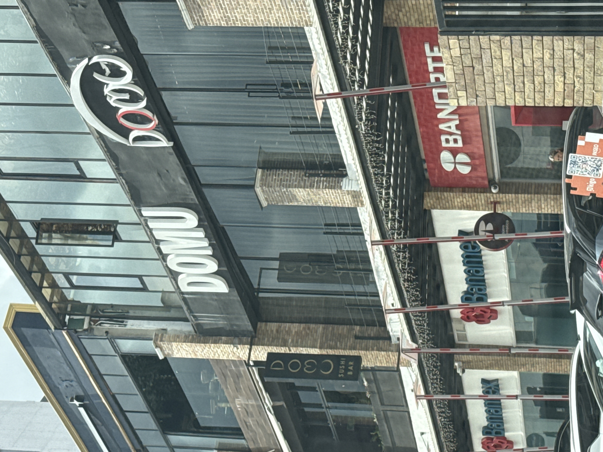 The image shows a street in what appears to be a commercial district. There are several businesses visible, including "DOMU" with a distinctive logo, "C300 Sushi Bar", a Banorte bank, and a Banamex branch. A black car is on the right side of the frame, equipped with a QR code sticker on the rear windshield. A security barrier is in place. The reflection in the glass windows suggests other buildings are across the street. This scene appears to be in Puebla, Mexico, due to the presence of Banorte and Banamex banks, which are commonly found in Mexico.