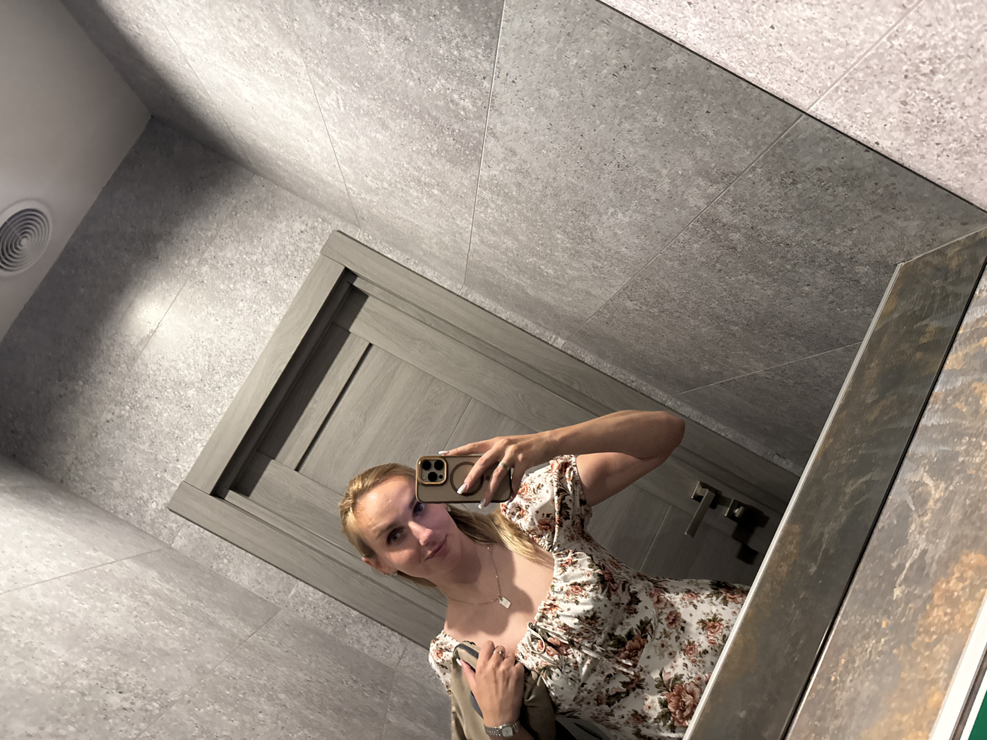 A woman with blonde hair is taking a mirror selfie in a modern restroom. She is wearing a white floral print dress with puff sleeves and a necklace. In her right hand, she holds a smartphone with a gold case, and her left hand is resting on her bag. The restroom features large grey tiles on the walls and floor, a wooden door, and a circular ventilation grate on the wall. The lighting suggests it is daytime. The woman has a slight smile and appears to be in a casual setting.