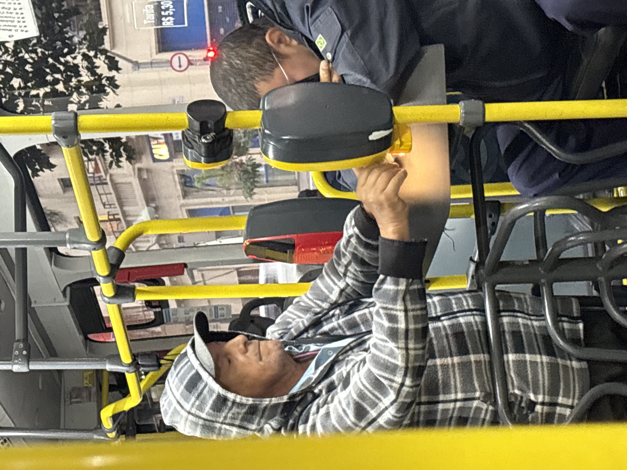 The image depicts the interior of a public bus in São Paulo, Brazil. Two men are visible. The man in the foreground, on the left, wears a plaid hooded jacket and a baseball cap. He appears to be in his 60s, with fair skin and receding hairline. His right hand is extended, interacting with a yellow and gray device attached to a yellow pole. The man in the background, on the right, wears a dark blue uniform or jacket and glasses. He is leaning forward, his head resting on his arms, and his face is obscured. The bus interior features bright yellow poles and gray railings, common in São Paulo's public transport system. Outside the bus window, elements of an urban environment are visible, including buildings and trees. A sign displaying "Tarifa R$ 5,30" indicates the fare in Brazilian Reais, confirming the location in Brazil. The overall lighting suggests it is daytime.