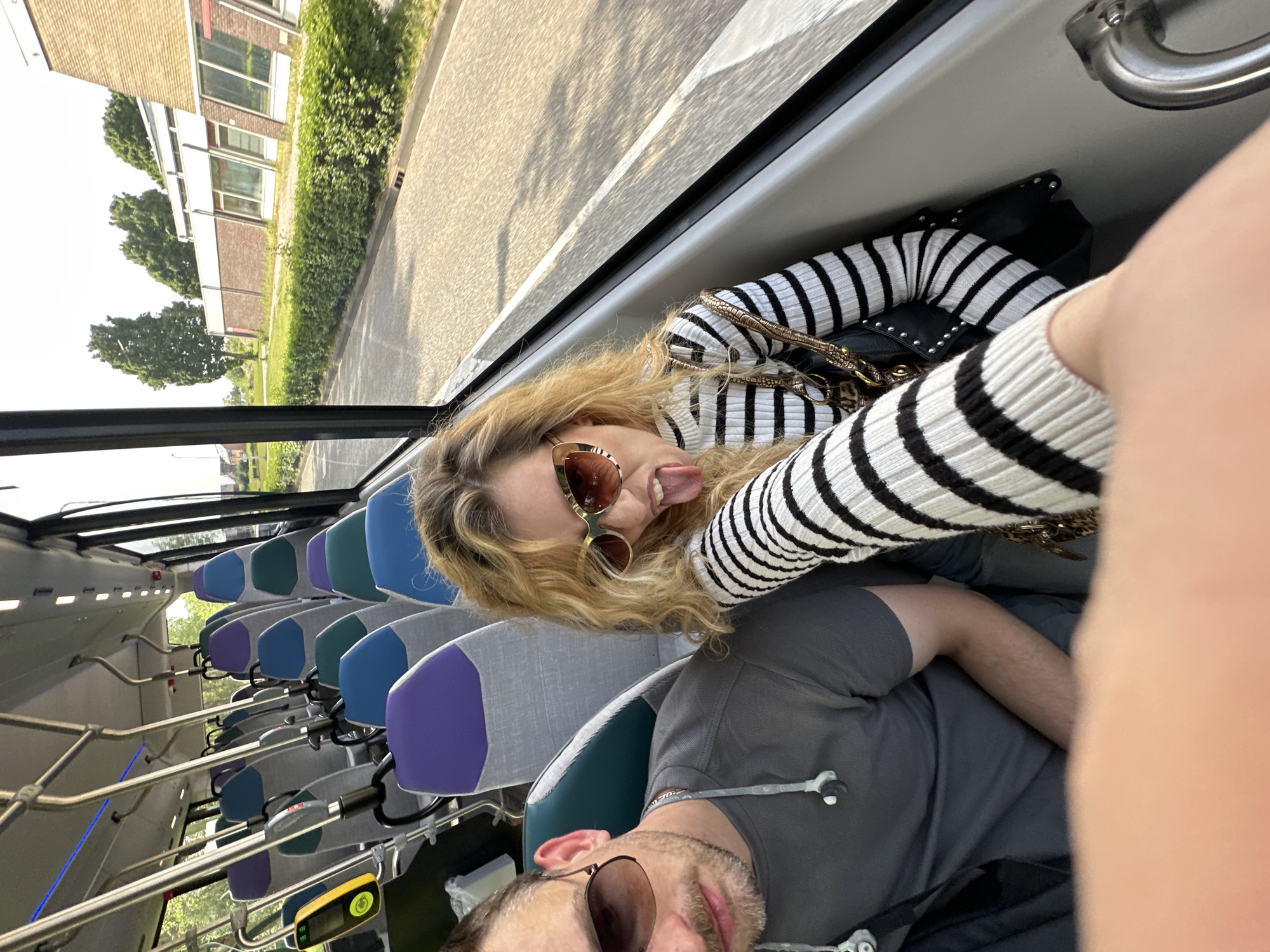 Inside a bus in Dronten, Netherlands, a woman with blond hair and sunglasses, wearing a black and white striped sweater, sticks her tongue out towards the camera. Beside her, a man with sunglasses and a gray shirt is visible. The interior of the bus features rows of seats with alternating blue and purple headrests. Outside the bus window, buildings, trees, and a street are visible, suggesting a cityscape.