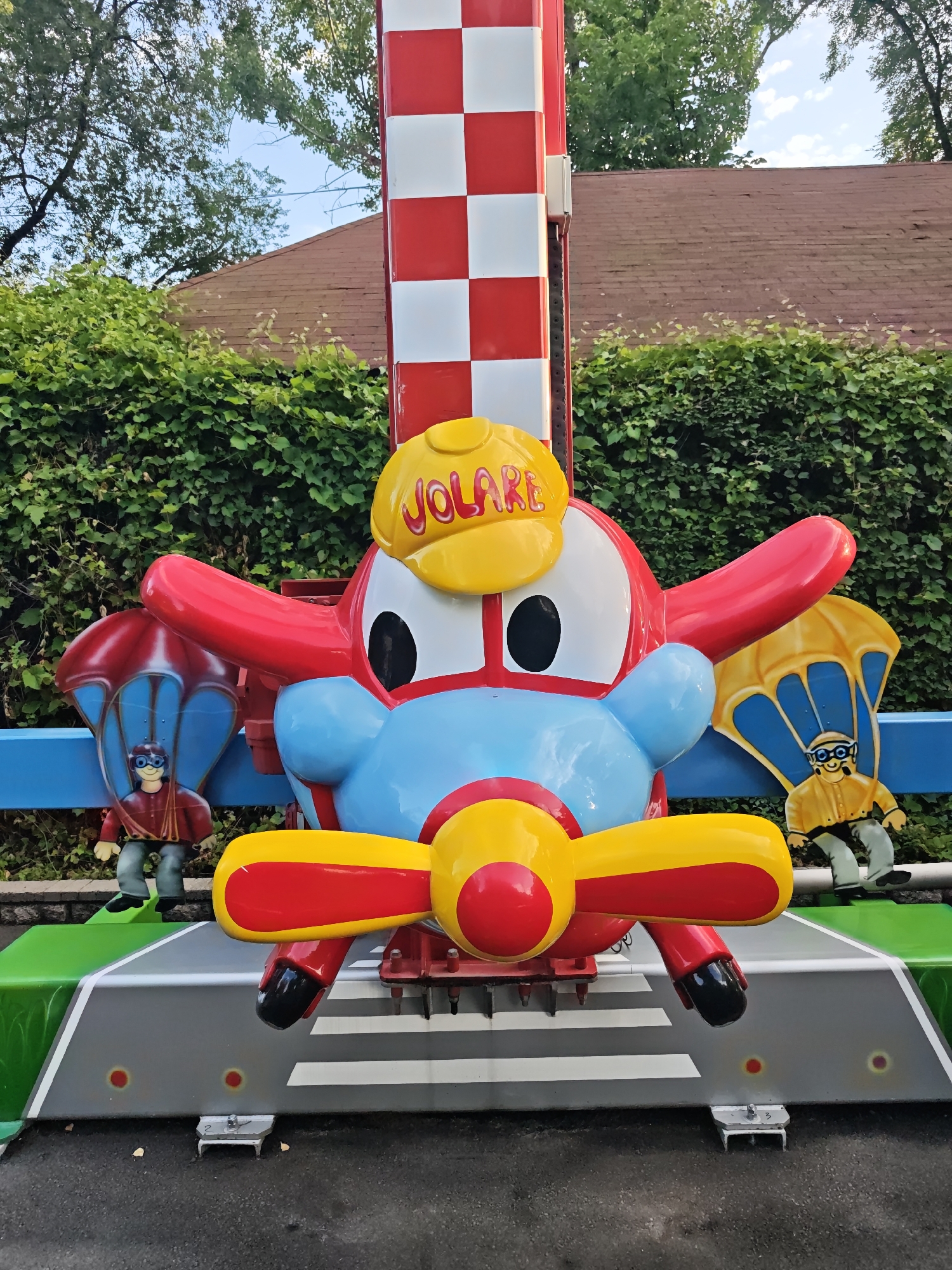 An amusement park ride features a cartoon airplane with red wings, a blue body, and a yellow propeller. The airplane has large white eyes with black pupils and a yellow baseball cap on its head with the word "JOLARE" printed in red. Two figures in pilot costumes are positioned on either side of the airplane. The figure on the left wears a red jacket and blue pants, holding a blue parachute. The figure on the right wears a yellow jacket and gray pants, holding a yellow parachute. The ride's structure is a tall, red and white checkered pole. The ride is set against a backdrop of dense green foliage and a brown-roofed building. The ride's base is painted green and gray, resembling a runway with white stripes. The scene appears to be outdoors during daylight.