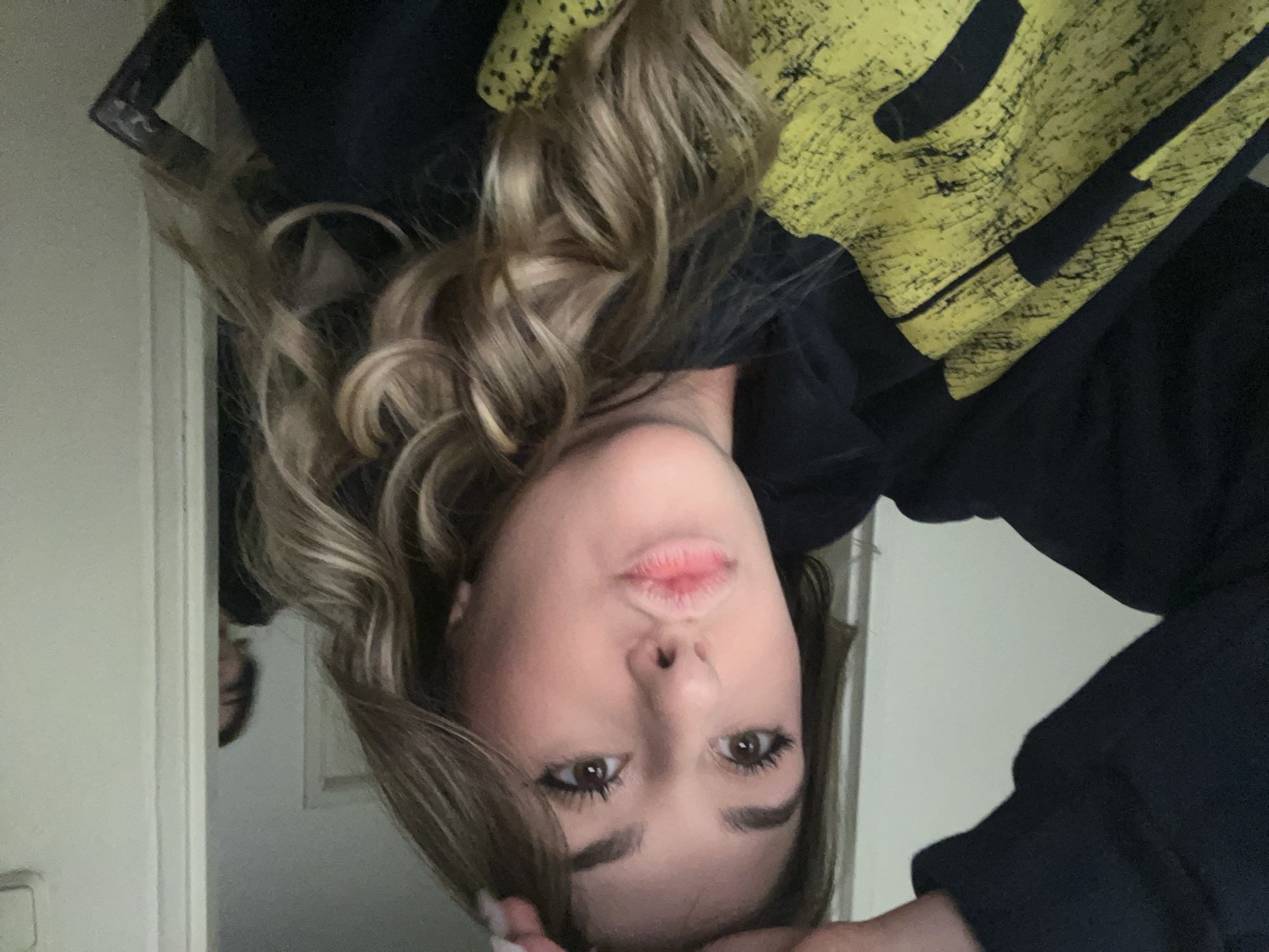 A young woman is holding the camera upside down in a portrait selfie. She has long blonde and light brown wavy hair, brown eyes, and pink lips. She is wearing a black hoodie with a large yellow design on the upper right side. A white door is visible behind her on the left. In the background, a blurry figure is visible through the partially open door. The location is not discernible as Sofia, Bulgaria based solely on the visual information provided in the image.