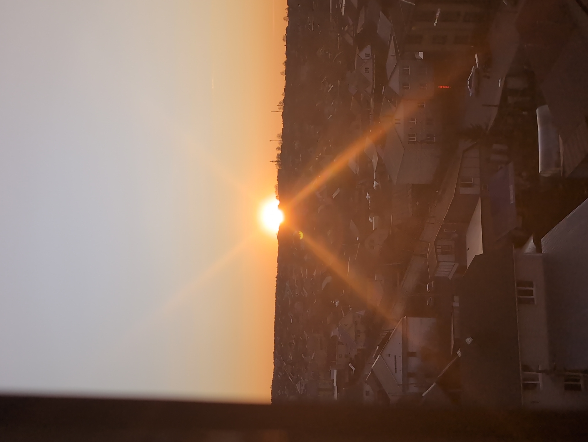 The image shows a sunset over the city of Izhevsk, Russia. The sun is low on the horizon, creating an orange glow in the sky. Silhouettes of buildings and trees are visible in the foreground. The image is taken from a high vantage point.