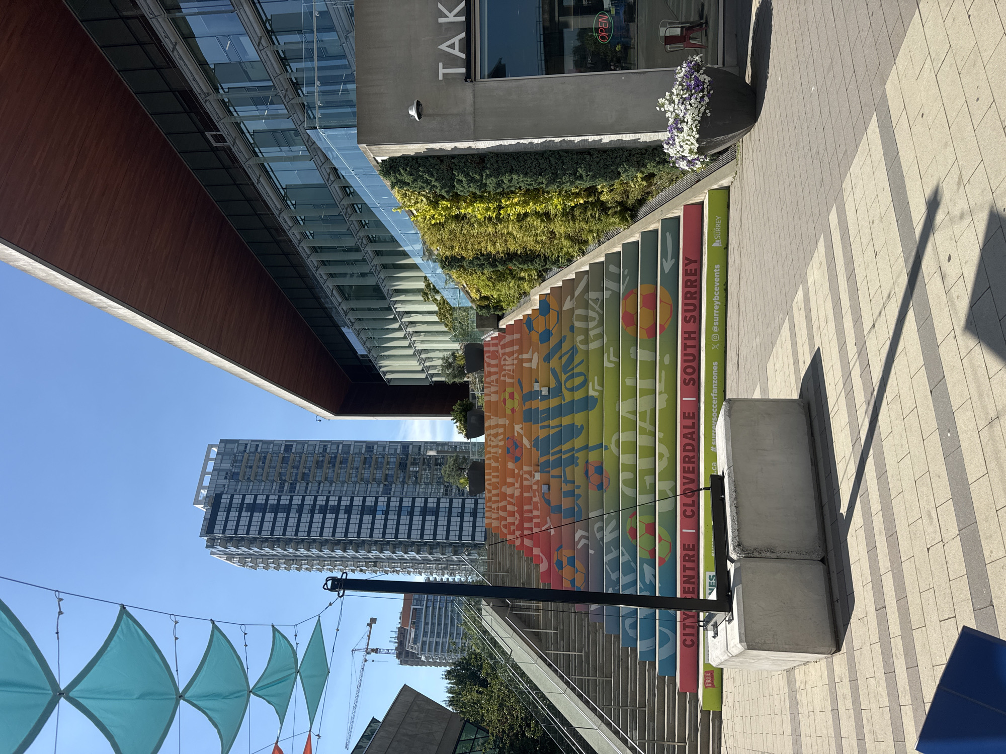 This image captures a vibrant, outdoor scene in Surrey, Canada, likely in a public plaza or park area. The focal point is a wide staircase adorned with a colorful mural that spans across its risers. The mural features large, bold text and graphic elements, including soccer balls, in a rainbow gradient of colors such as red, orange, yellow, green, and blue. The text includes words and phrases like "GOAL," "CHILL," "WATCH PARTY," "CITY CENTRE," "CLOVERDALE," and "SOUTH SURREY," suggesting a connection to local community events or sports.
The staircase leads up towards a modern, multi-story building with extensive glass facades and a distinctive wooden overhang. To the left of the stairs, a contemporary skyscraper with numerous windows rises against a clear blue sky. Several teal-colored triangular fabric shades are suspended overhead, casting dappled shadows.
Adjacent to the building, a vertical garden with lush green foliage adds a natural element to the urban landscape. A dark gray planter filled with white and purple flowers sits near the entrance of the building. The ground in front of the stairs is paved with a patterned walkway of light and dark gray rectangular stones. In the foreground, two large concrete blocks are positioned on the walkway, with a black metal pole rising from them. The overall environment appears clean, modern, and well-maintained, indicative of a public space designed for community gatherings or events.