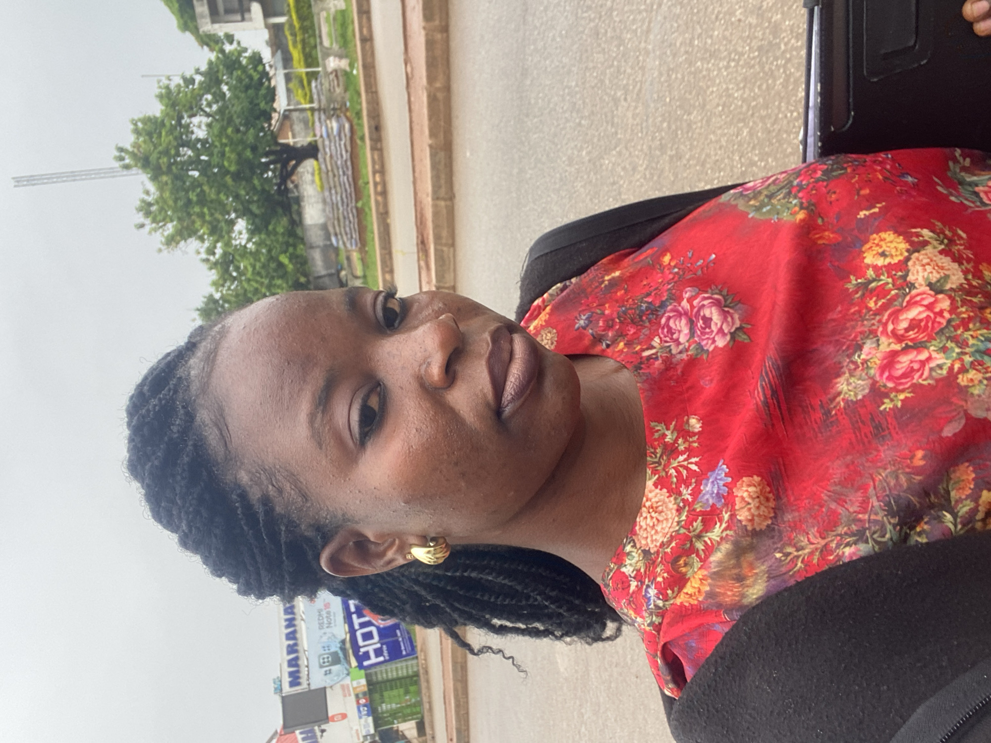 A woman with braided black hair and gold hoop earrings, wearing a red floral top and a black jacket, is pictured outdoors. The background suggests an urban setting in Bukuru, Nigeria, with a road, buildings, trees, and signage visible, including a billboard that says "MARANA" and "HOT". The sky is overcast. The woman is looking towards the camera with a neutral expression.