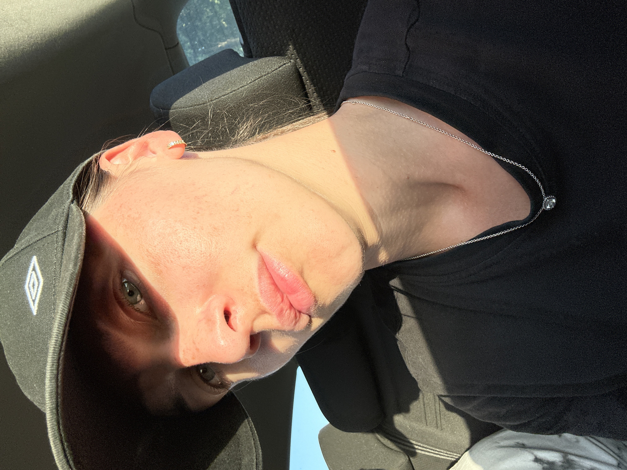 A close-up, slightly angled self-portrait depicts a fair-skinned person wearing a black baseball cap and a black T-shirt. A delicate silver necklace with a small, light-colored pendant rests on the T-shirt. The person has light-colored hair and is wearing a small silver earring in their upper ear. Light and shadow play across the face, obscuring part of the left eye under the shadow of the cap's brim. The lips are slightly parted. The background suggests an interior space, possibly a car, with a portion of a seat visible. There is no verifiable information to specify Bargstedt, Germany as the location.